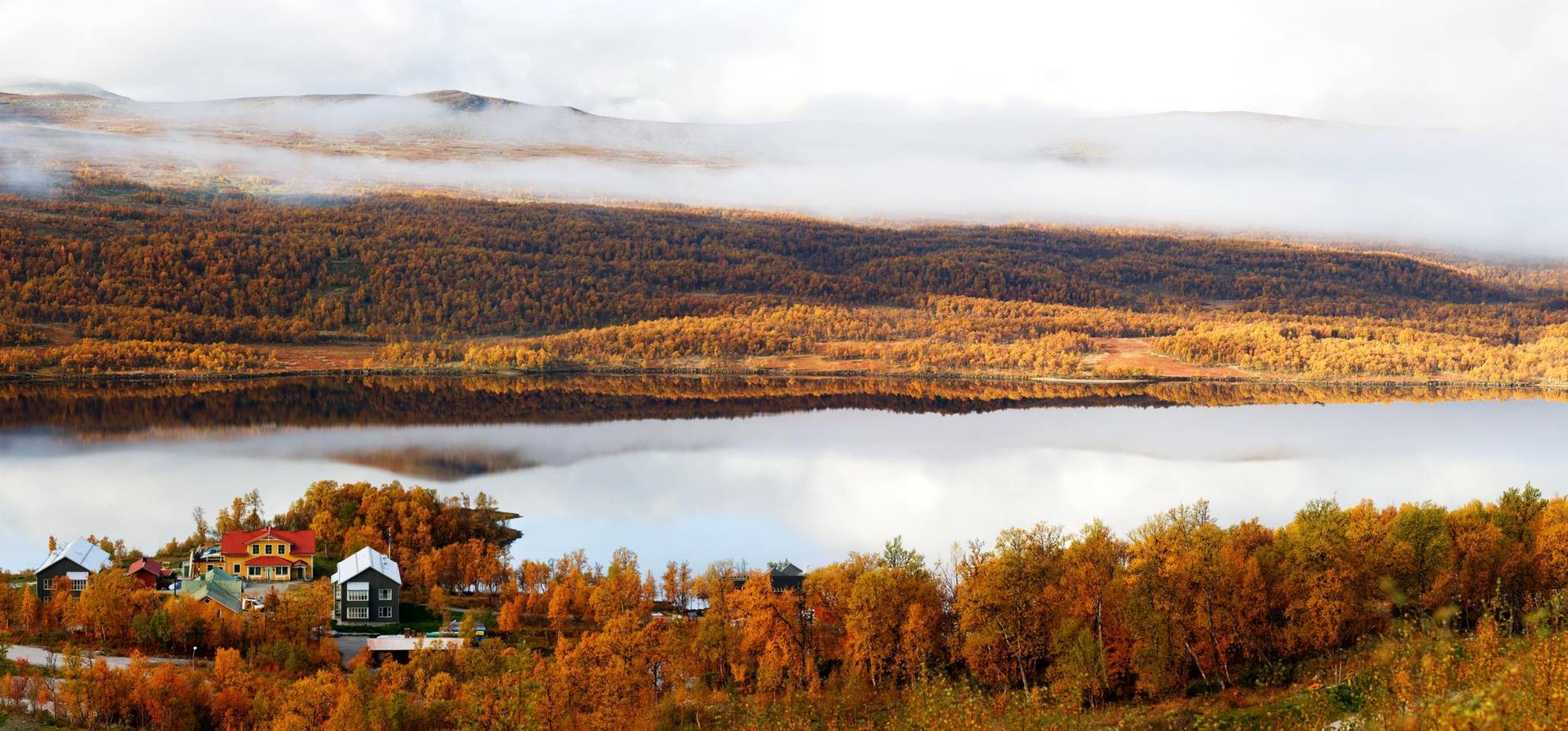 Une belle vue d'automne avec un hôtel au bord d'un lac dans les montagnes. Les arbres sont oranges et on aperçoit du brouillard qui s'étend, au-delà du lac, sur les montagnes