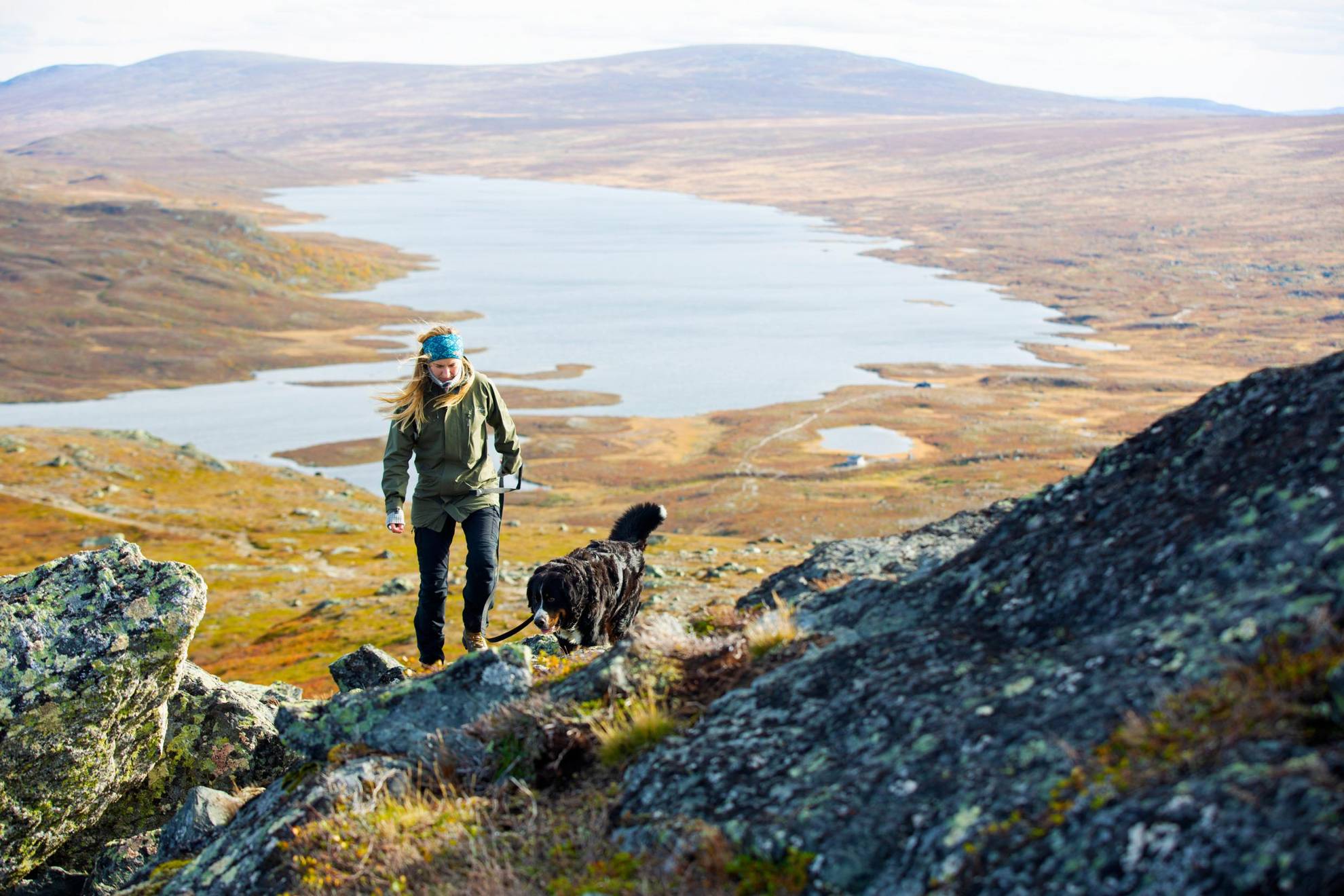 Une femme monte un versant de montagne avec son chien. En arrière-plan, il y a une vue panoramique et surplombante sur une vallée avec un lac au milieu.