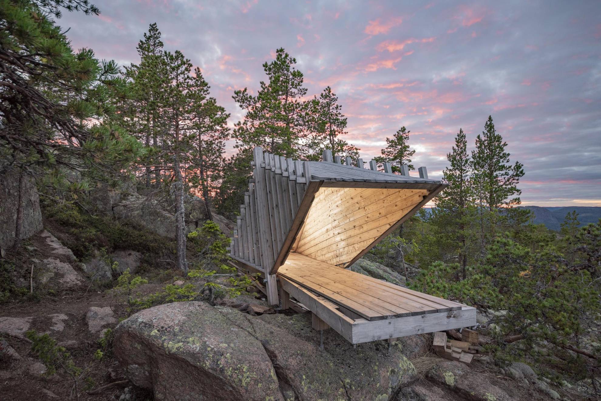 Une hutte ou une cabane construite avec une architecture minimaliste se dresse au sommet d'une colline boisée de la Haute Côte de Suède, avec le crépuscule derrière elle.