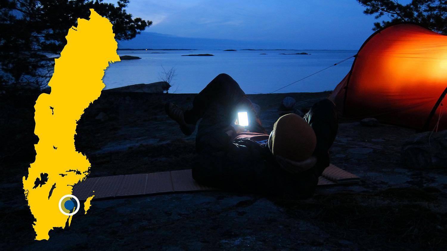 Une tente est plantée sur une plage près de l'eau. Il fait nuit et une personne est allongée à côté de la tente et regarde son téléphone.