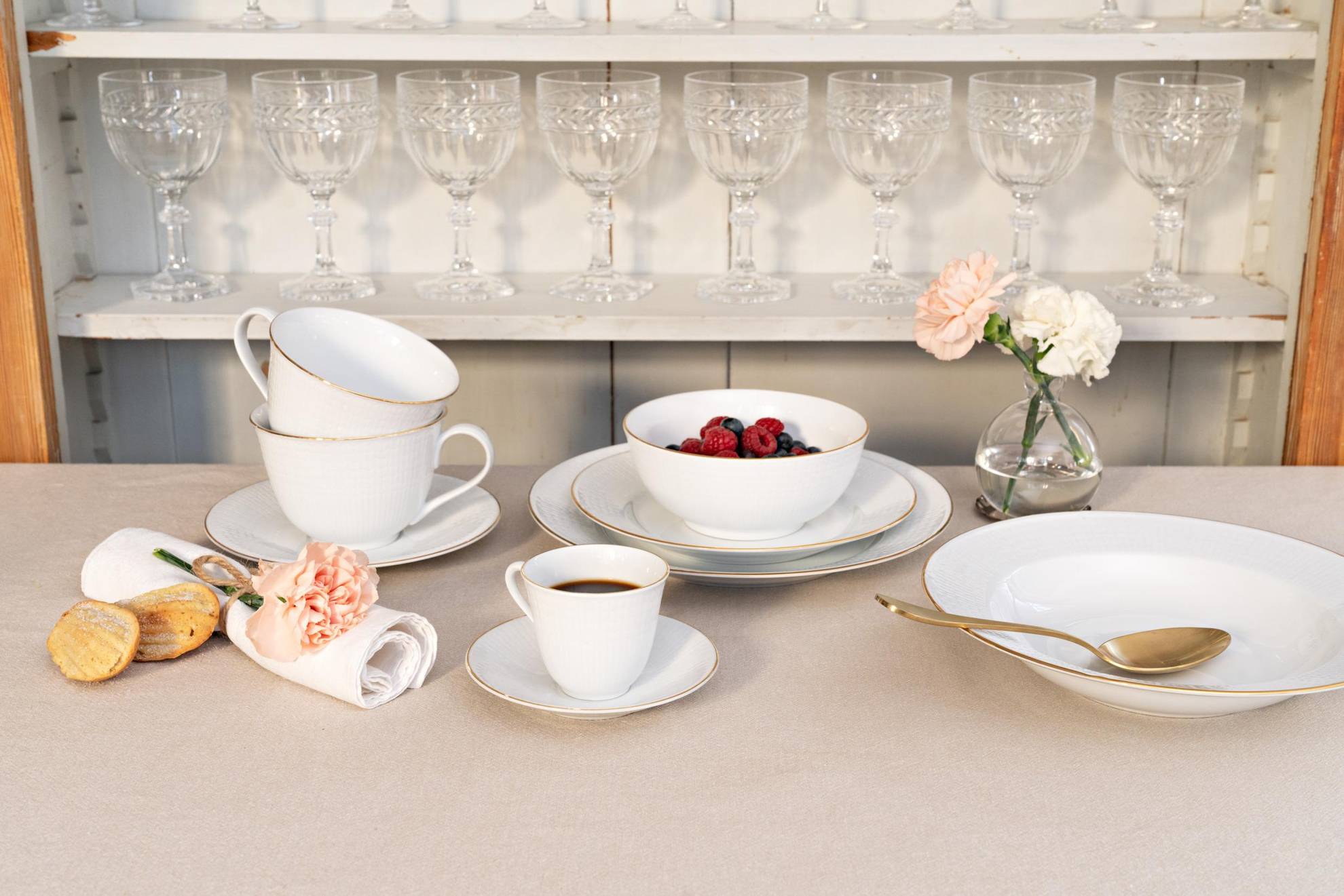Un service appelé "Swedish Grace Gala" est posé sur une table. Des assiettes de différentes tailles, un bol avec des baies et une tasse de café. Derrière, il y a une étagère avec des verres à vin.