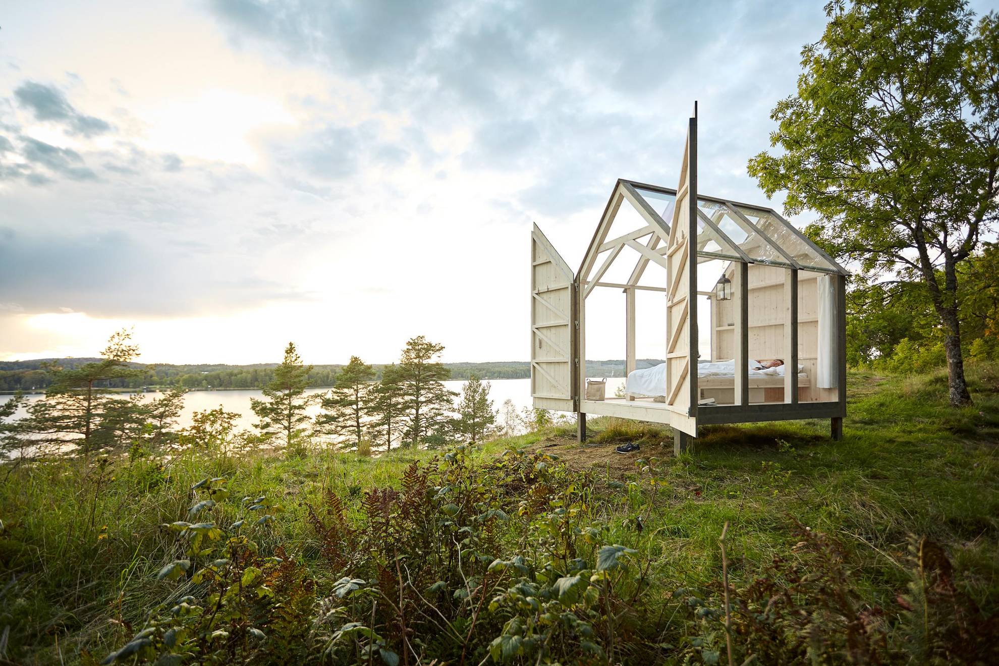 Une cabane en verre dans la nature avec les portes ouvertes. On voit au loin la mer et la forêt.