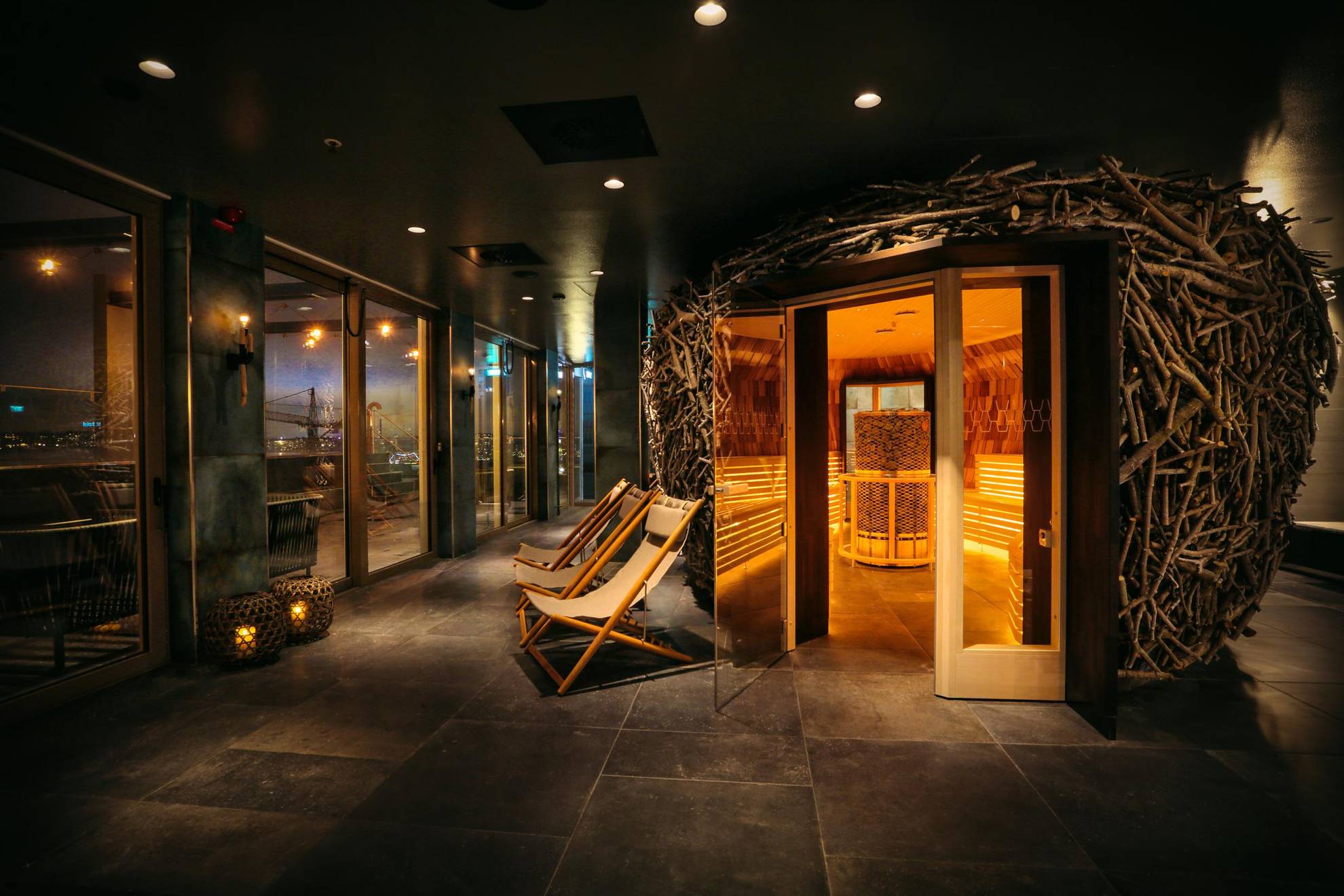 Un sauna ressemblant à un nid d'oiseau est situé au centre d'une pièce peu éclairée. On peut voir trois chaises longues juste à côté du sauna.