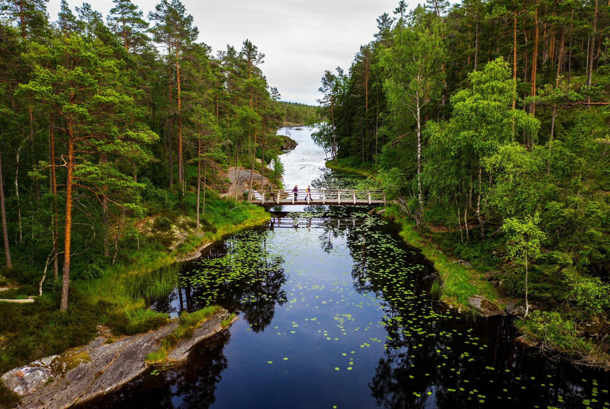 Trois personnes marchent sur un pont en bois étroit au-dessus d'un lac. La forêt borde les deux rives du lac.