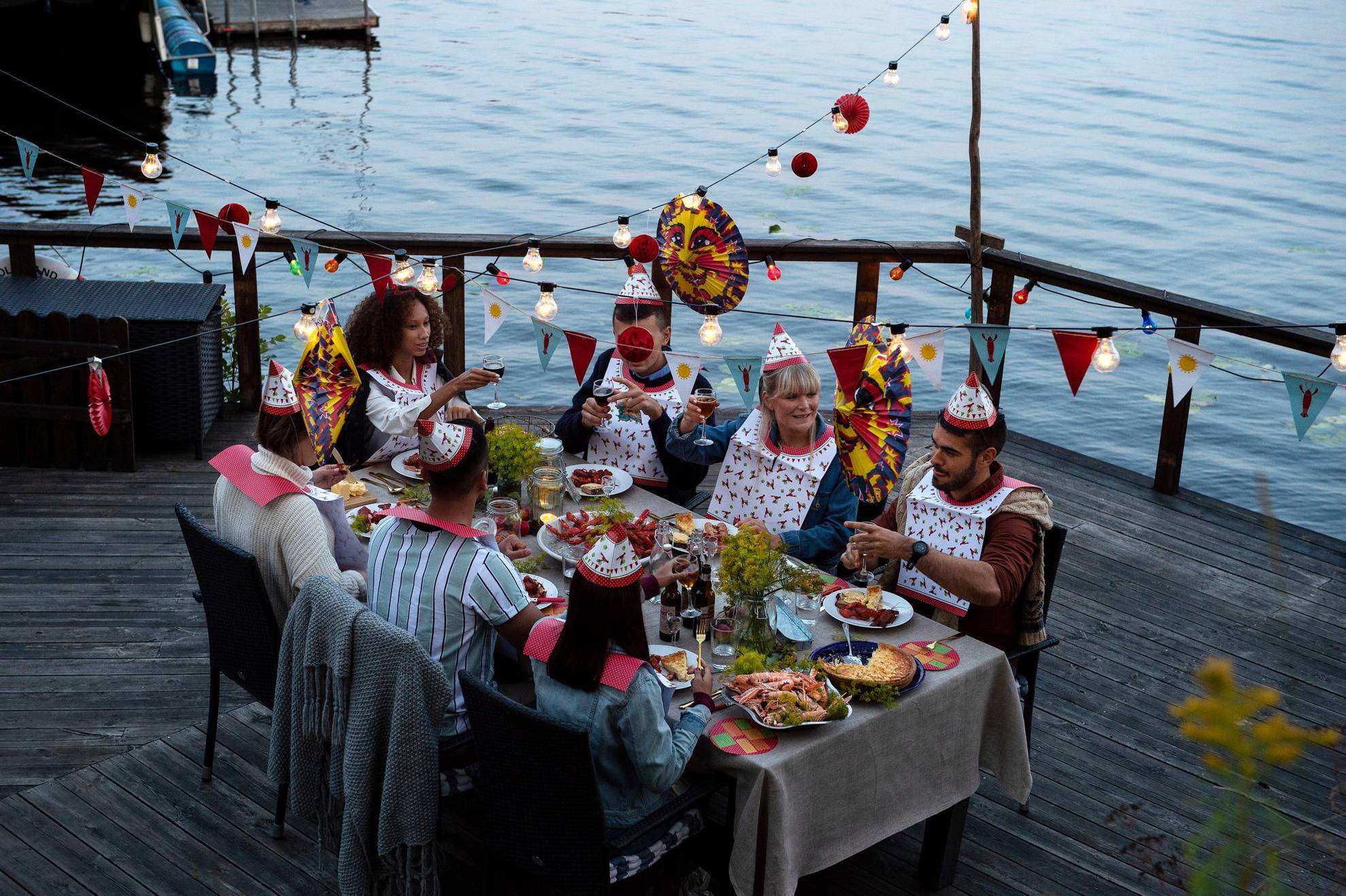 Une fête des écrevisses sur un ponton au bord de l’eau. Sept personnes sont assises autour d'une table, vêtues de chapeaux et serviettes en papier accrochées autour du cou et mangent des écrevisses. Au-dessus de la table sont accrochées des lanternes en papier, des lumières et des décorations.