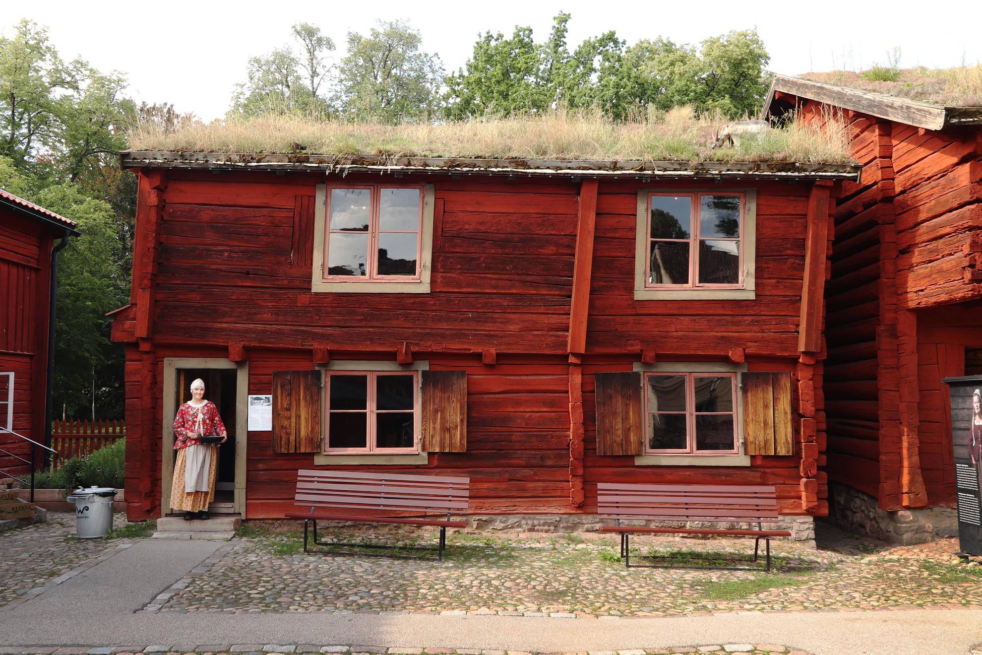 Une ancienne maison en bois peinte en rouge avec un toit en terre. Une femme se tient debout au niveau de l'entrée de la maison.