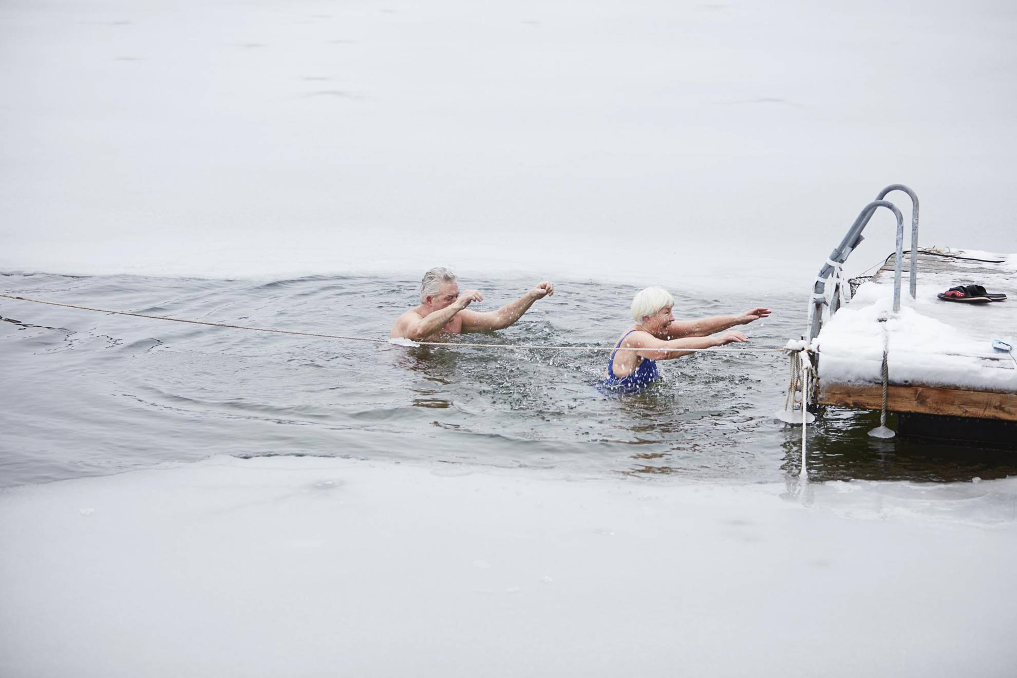 Un couple de personnes âgées se baignent dans un trou réalisé dans un lac recouvert de glace. Ils sont en train de retourner vers l'échelle sur la jetée.