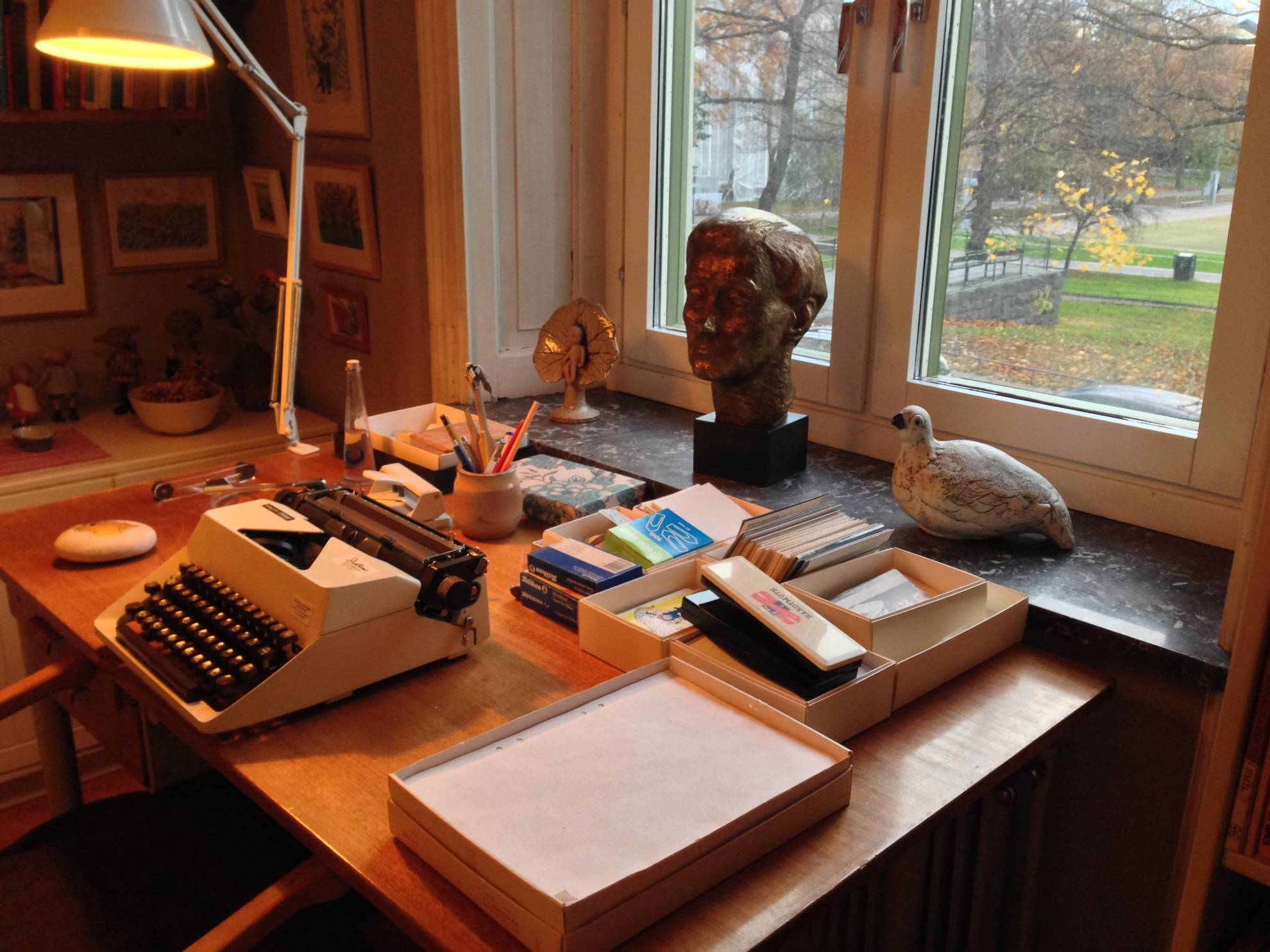 Author Astrid Lindgren's desk in Stockholm, filled with a typewriter, sheets of paper, a reading lamp and some decorations, with a view of a back garden.