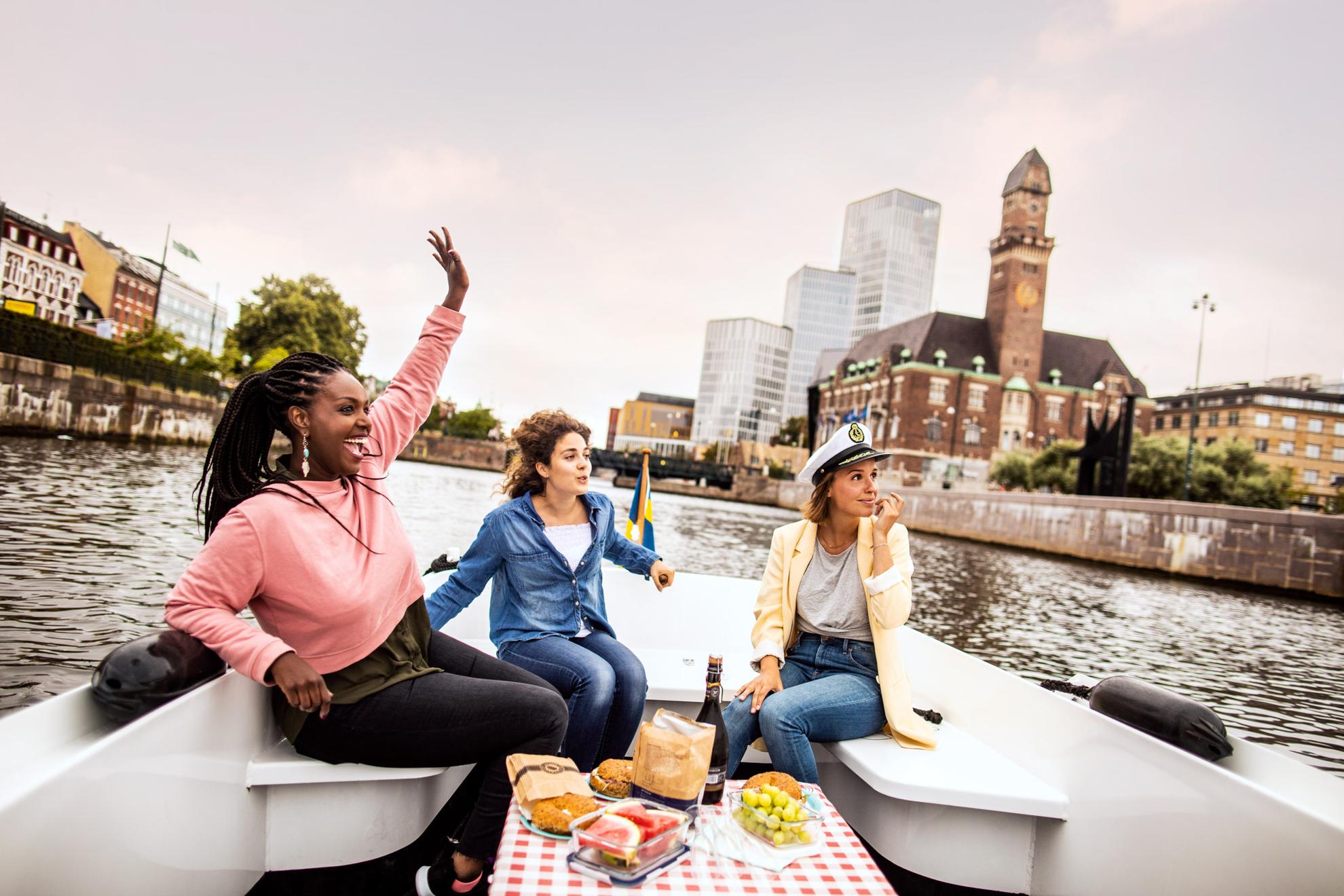 Trois jeunes femmes heureuses en excursion dans un bateau sur le canal de Malmö, avec Malmö Live et la gare centrale en arrière-plan.