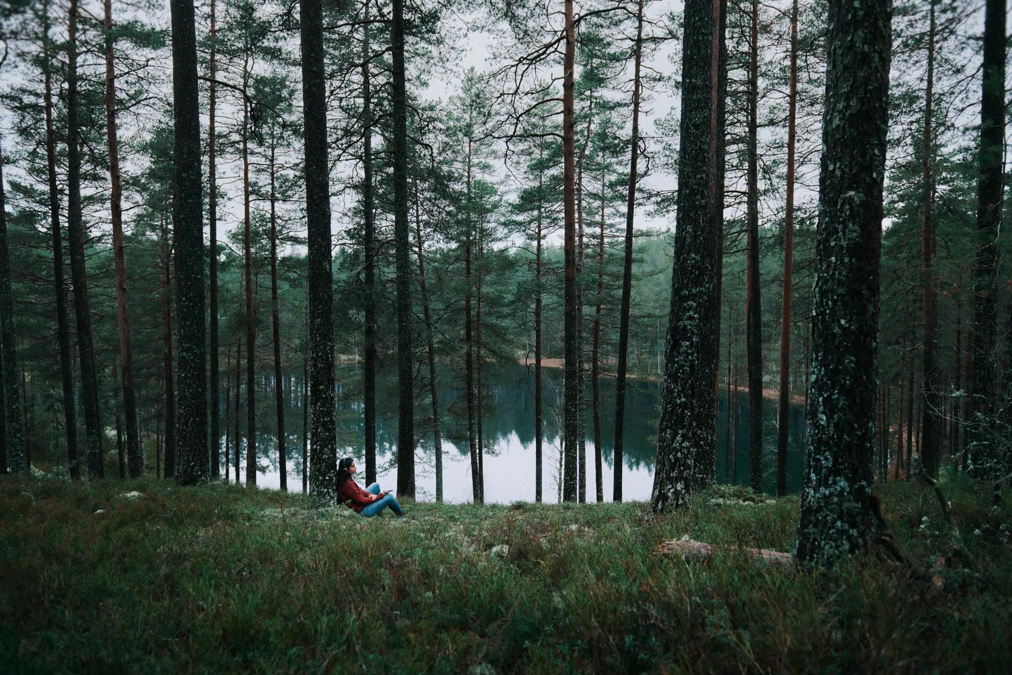 Une femme portant des écouteurs se repose contre un arbre à côté d'un lac. Le temps est maussade.