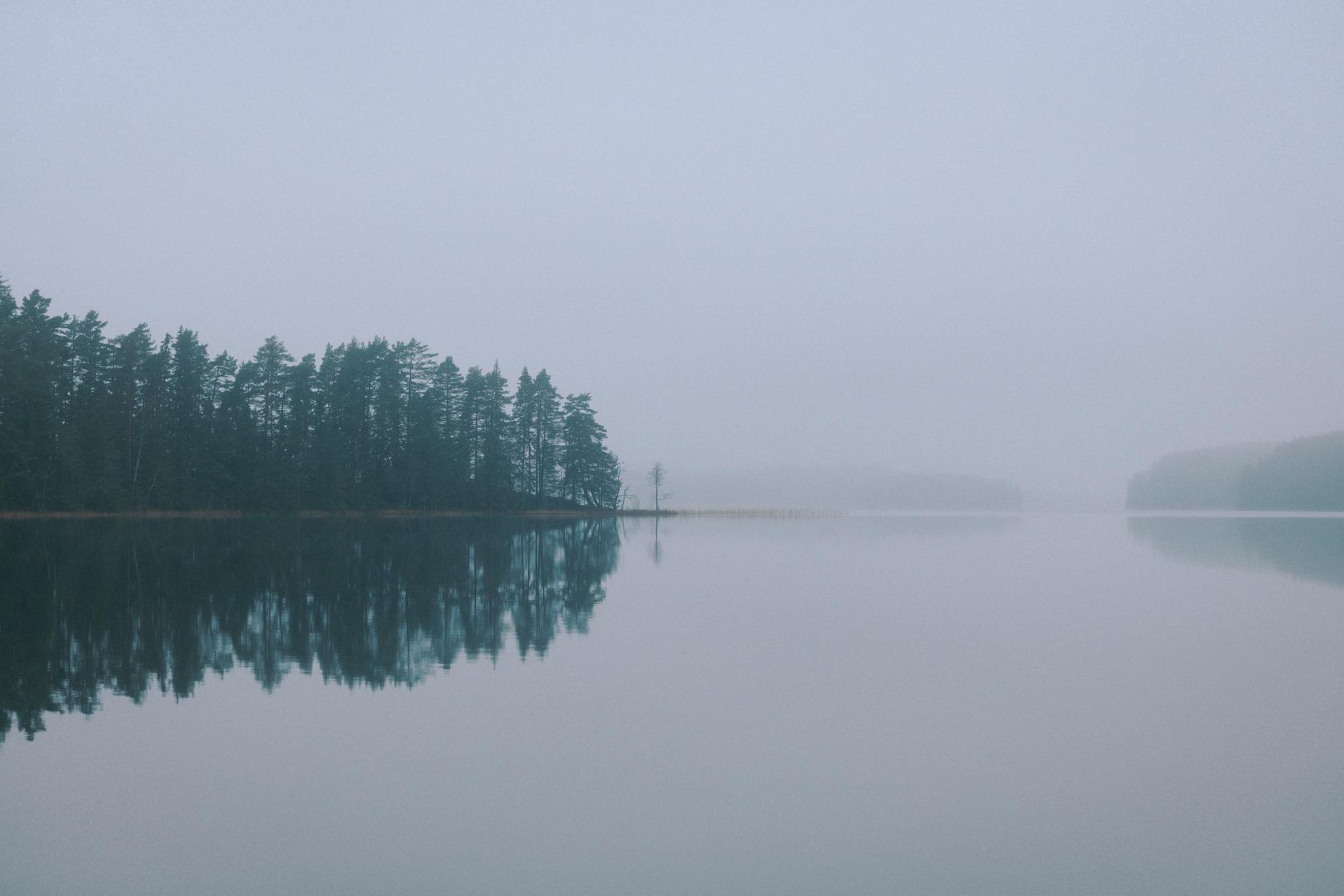 Un lac dans le brouillard. Les arbres verts et le ciel gris se reflètent sur l'eau calme.