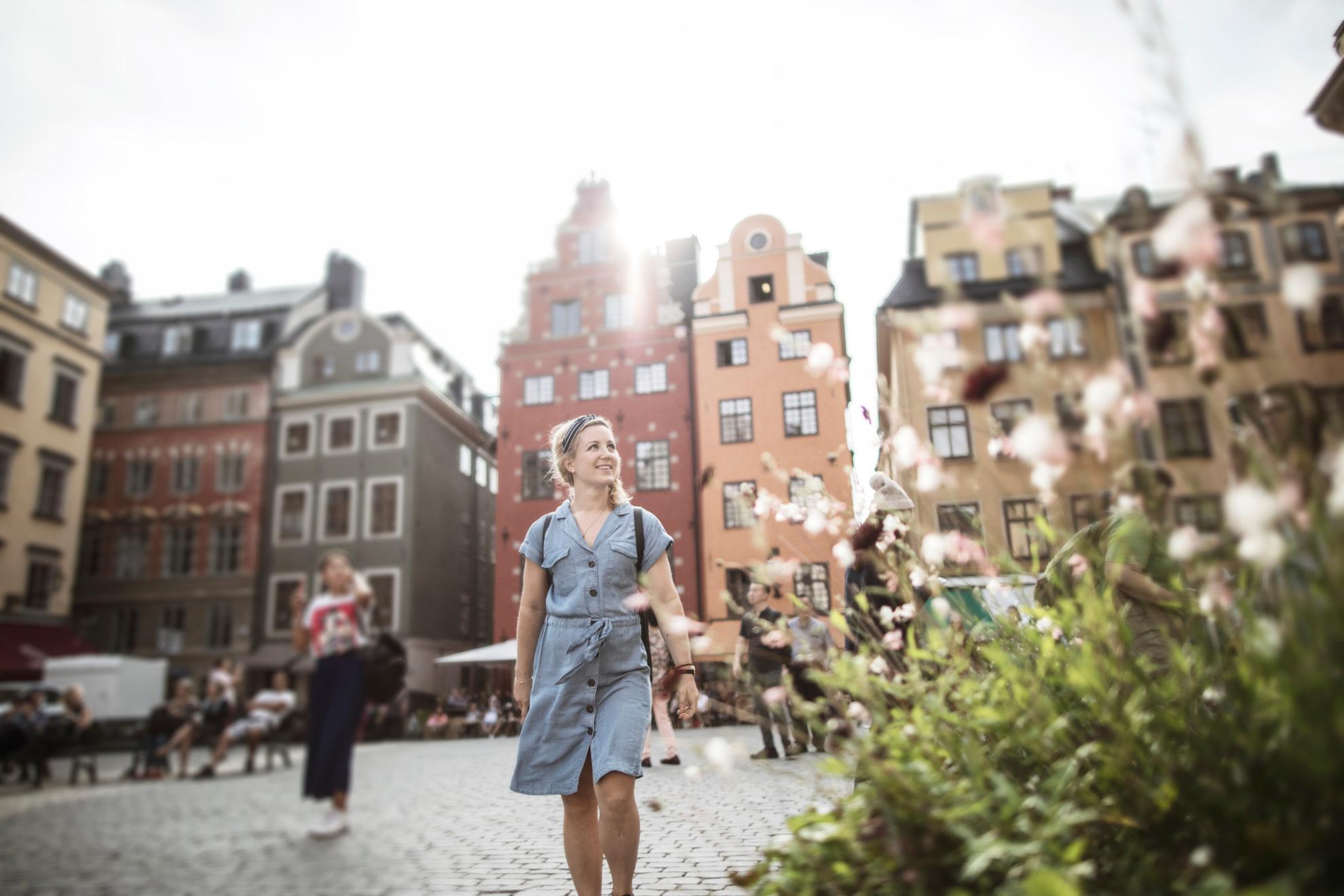 Une femme marchant sur une place de la vieille ville. À l'arrière-plan, on aperçoit de vieilles maisons en pierre et, à l'avant, une plante à fleurs.