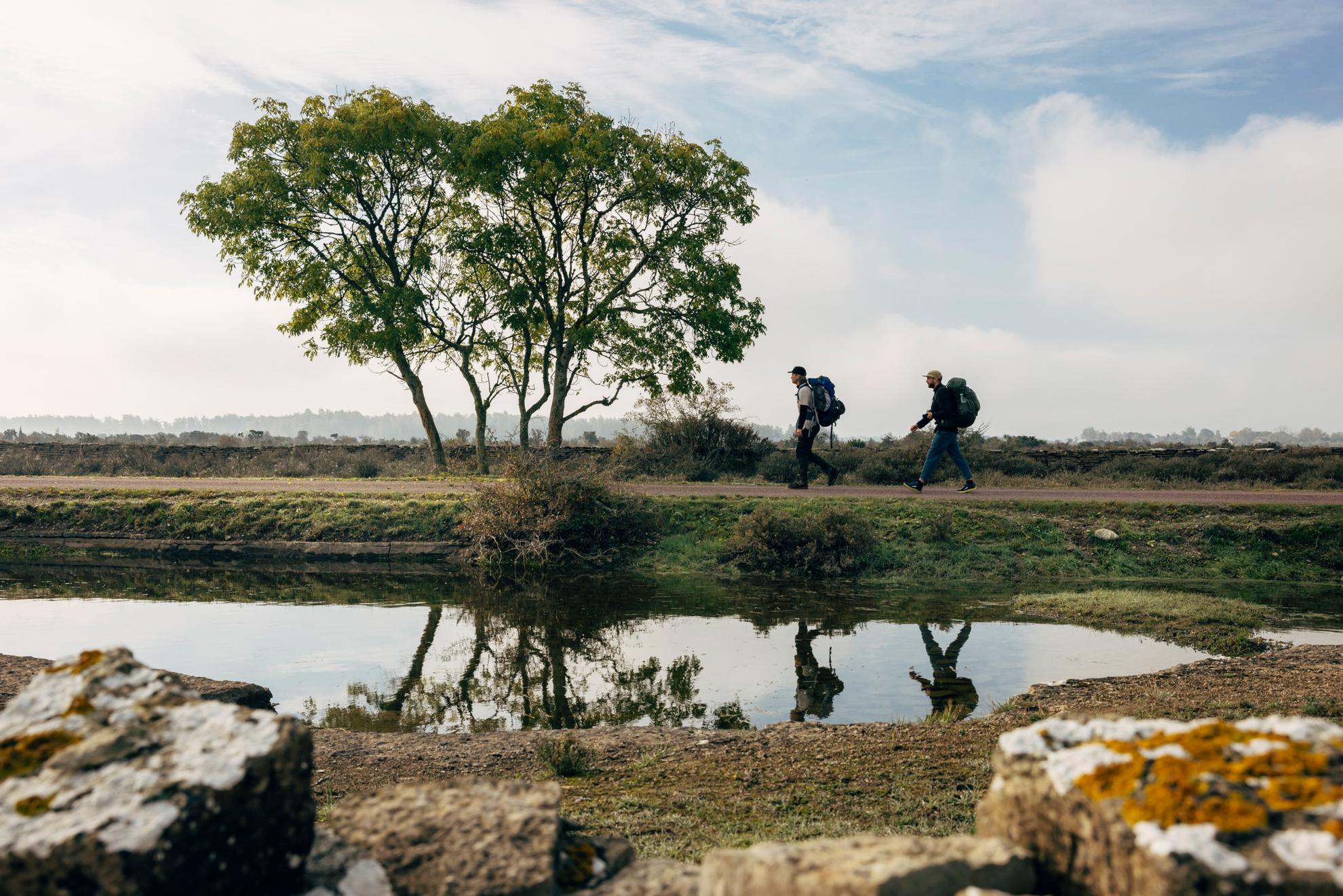 Deux randonneurs marchent près d'un petit étang avec des rochers au premier plan et des arbres à l'arrière-plan.