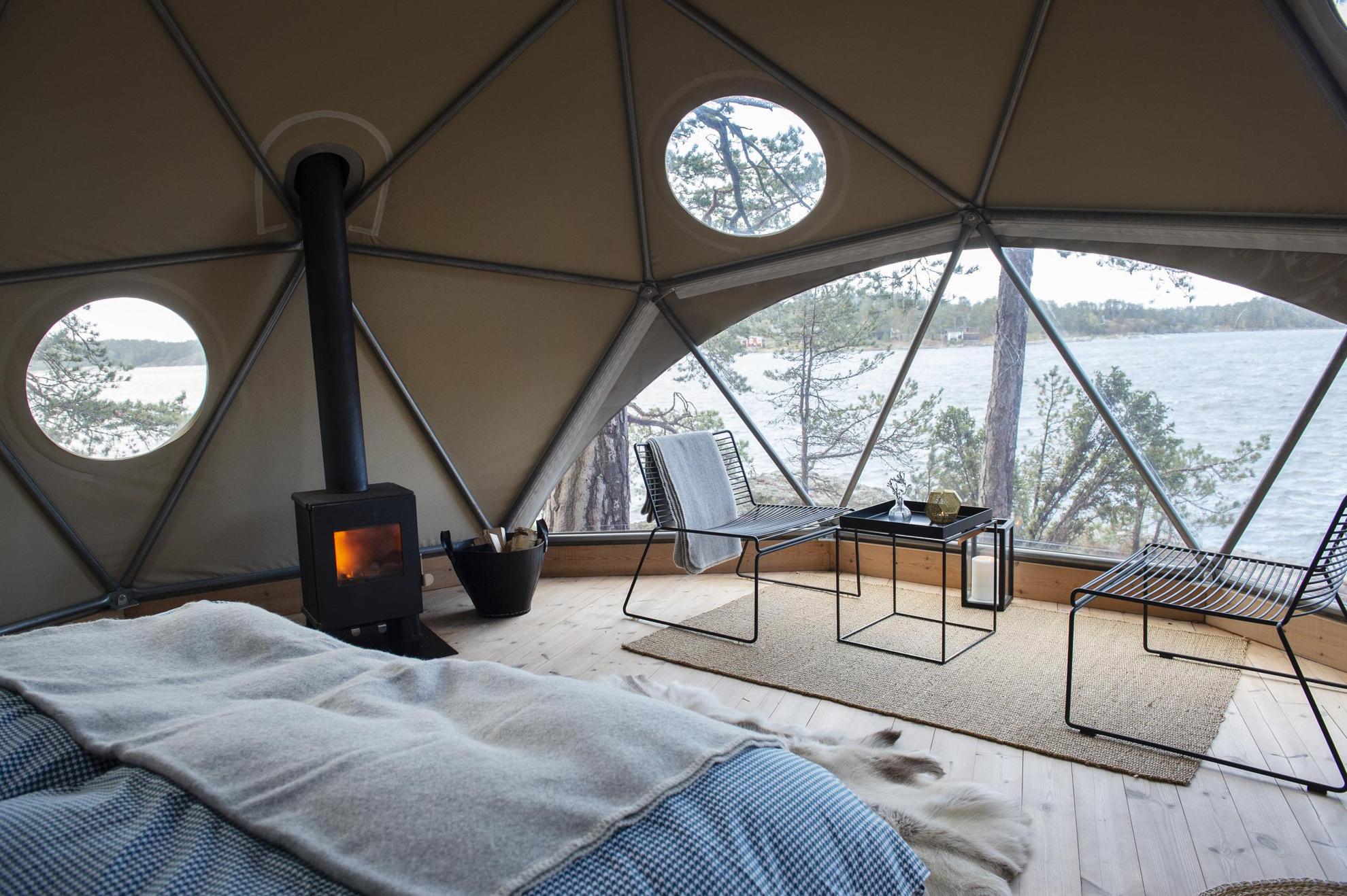 On voit l'intérieur d'une grande tente avec des chaises longues, une table basse, un lit, un poêle à bois. et en arrière-plan,  une vue sur la mer.