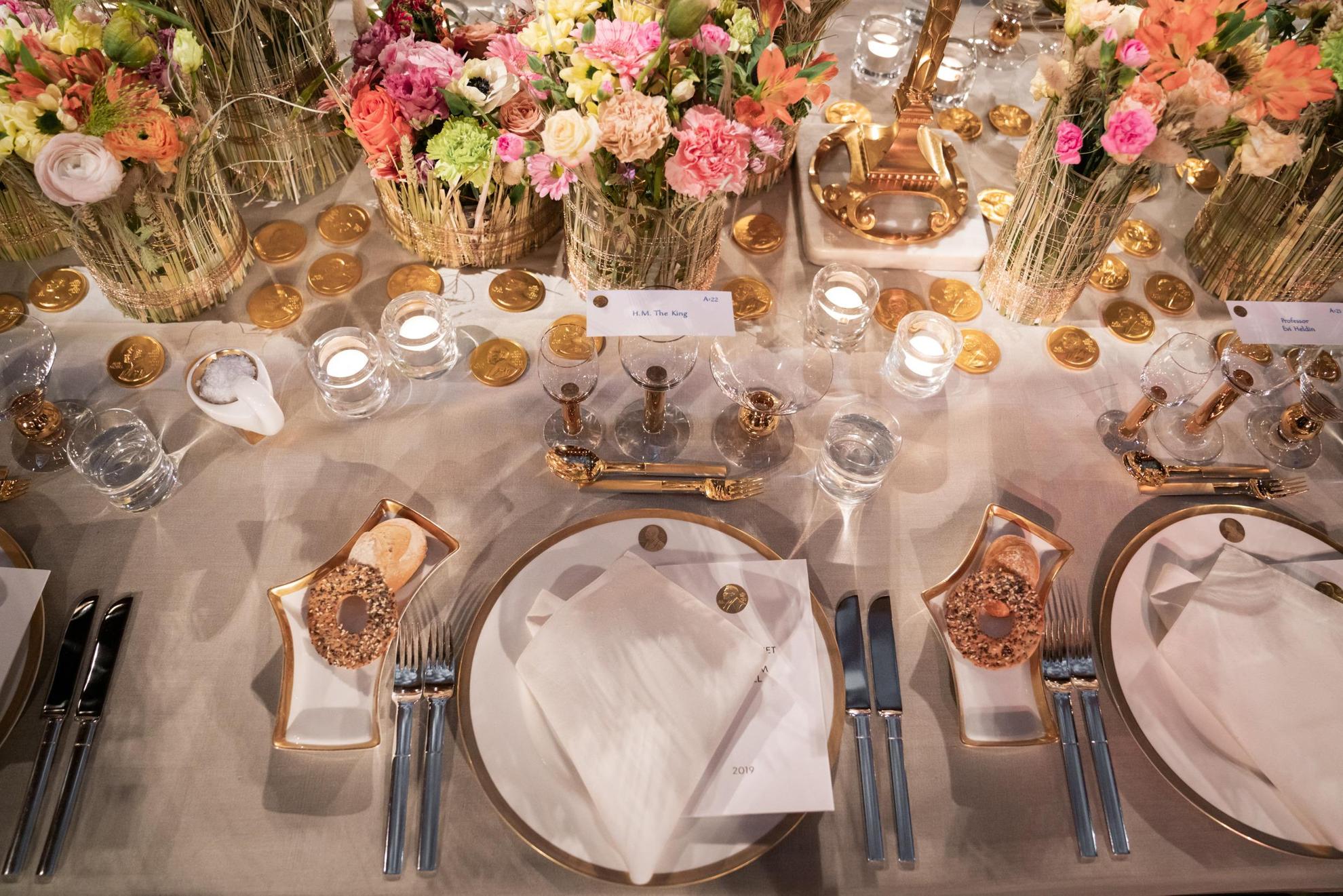 Une vaisselle blanche avec des détails dorés est posée sur une table avec une nappe blanche. Au milieu de la table se trouvent des fleurs colorées dans des vases.