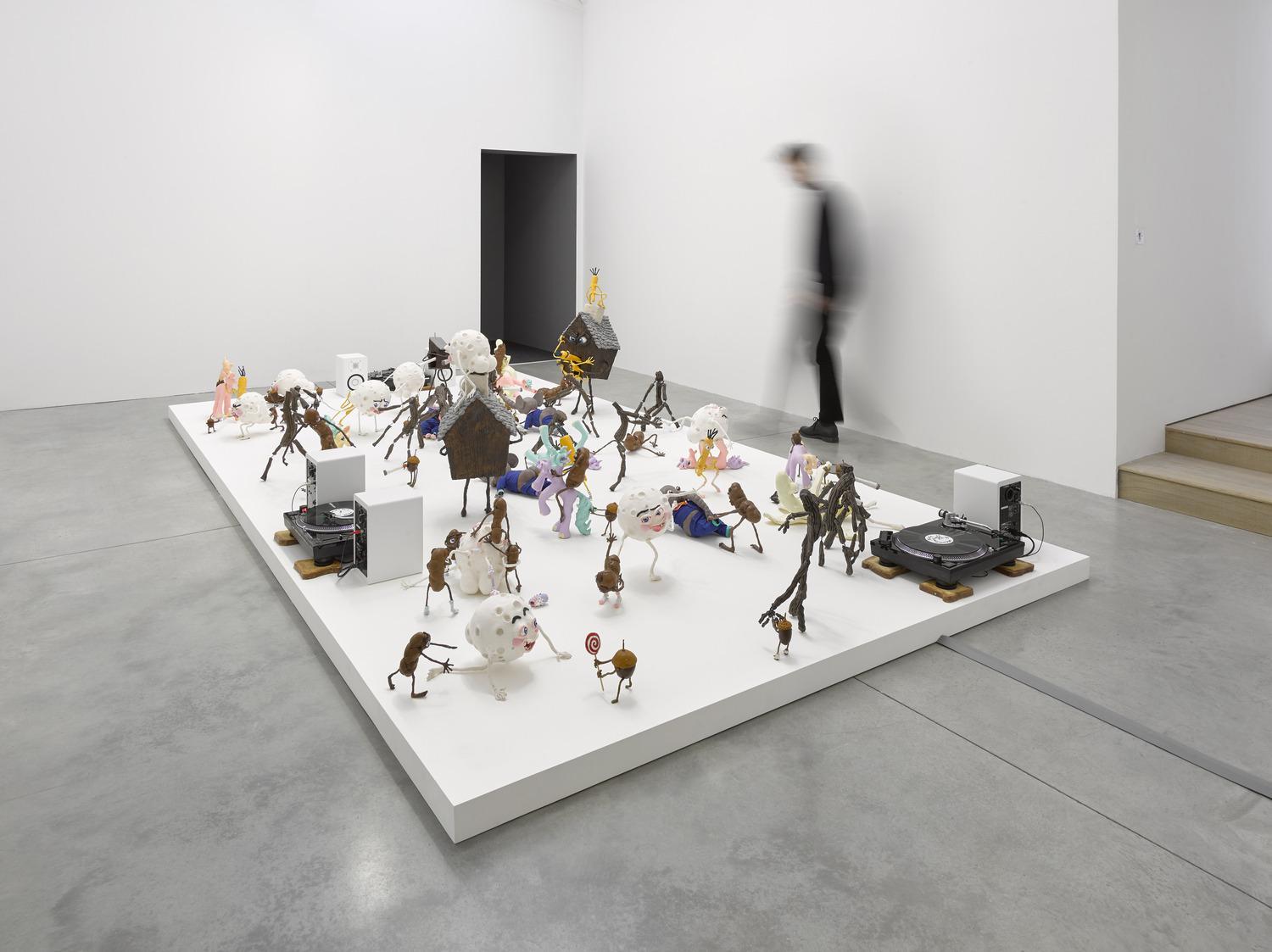 A spatial installation with several small sculptures and three record player, called ‘Who am I to Judge, or, It Must be Something Delicious, made by the artists Nathalie Djurberg and Hans Berg. A man is seen blurry in the background.