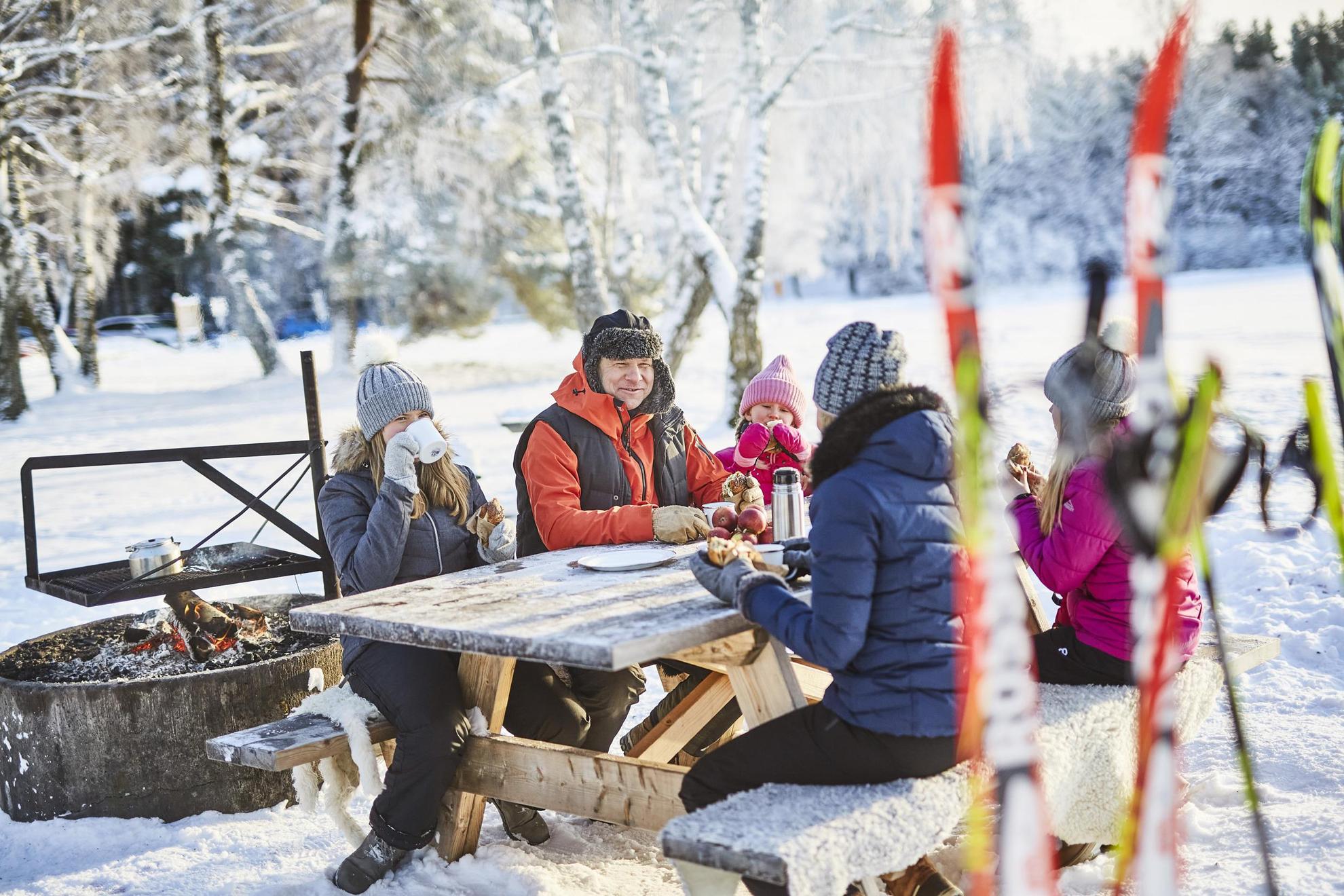 Une famille de cinq personnes assise dehors à une table, mangeant des brioches à la cannelle et buvant du café. Il y a des skis au premier plan de la photo.