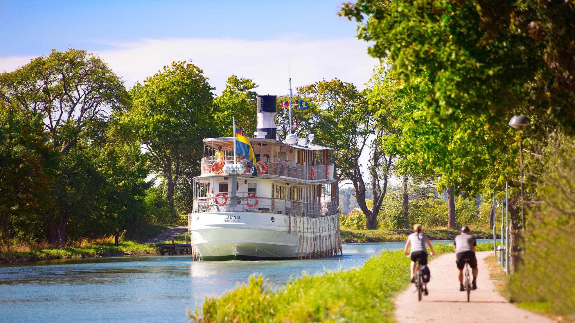C'est l'été et deux personnes font du vélo à côté de l'eau du canal Göta. Un bateau se déplace le long du canal.