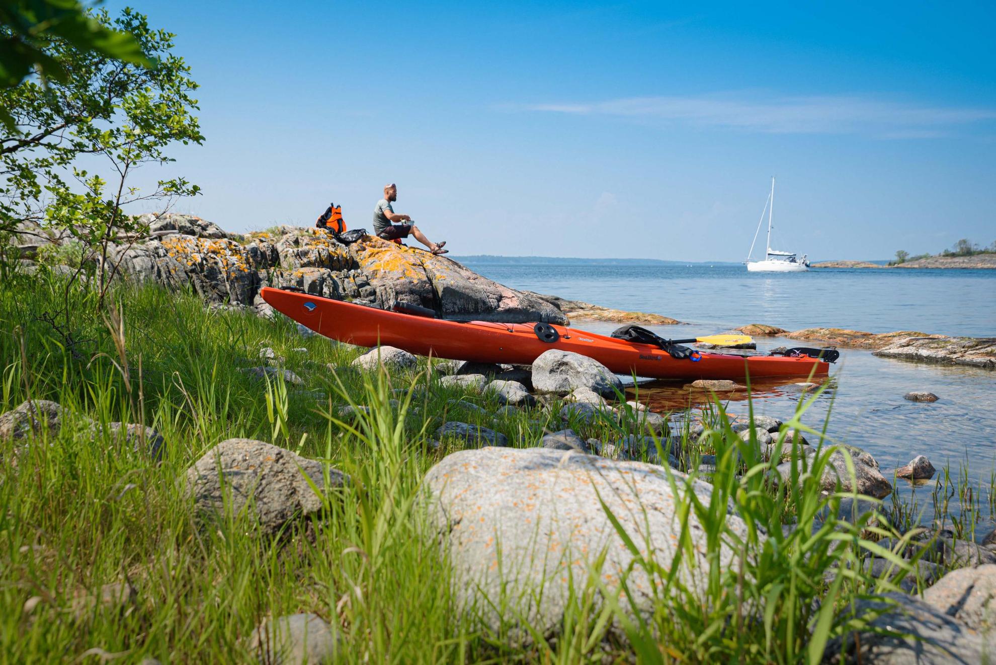 Un kayak orange est au bord de l'eau et un homme déjeune à côté sur un rocher. Un voilier blanc se trouve en arrière-plan.