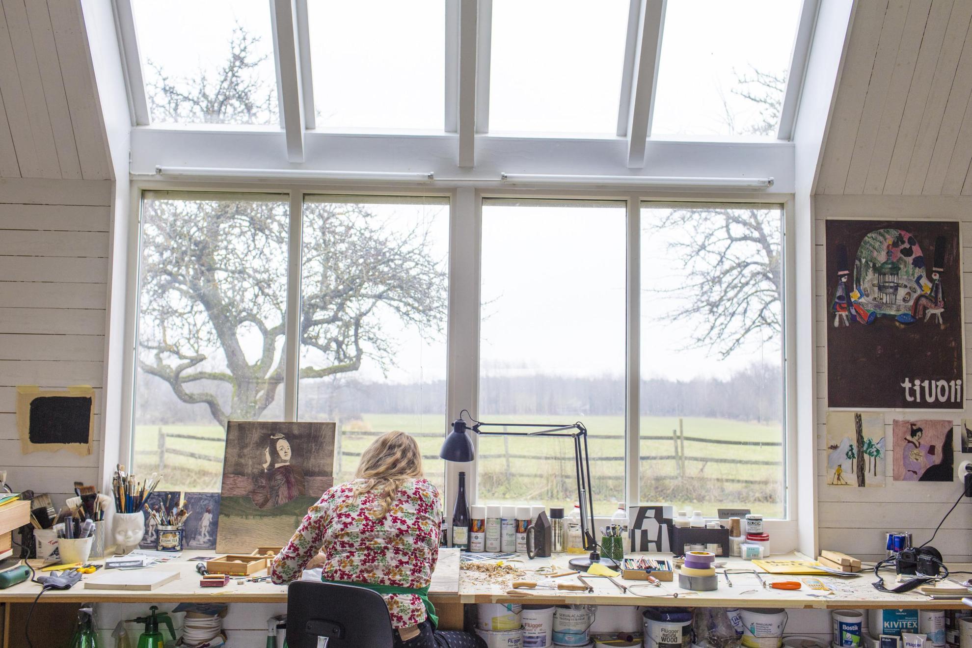 The artist Karin Mamma Andersson is seen from behind sitting at a table in front of large windows, in her studio.