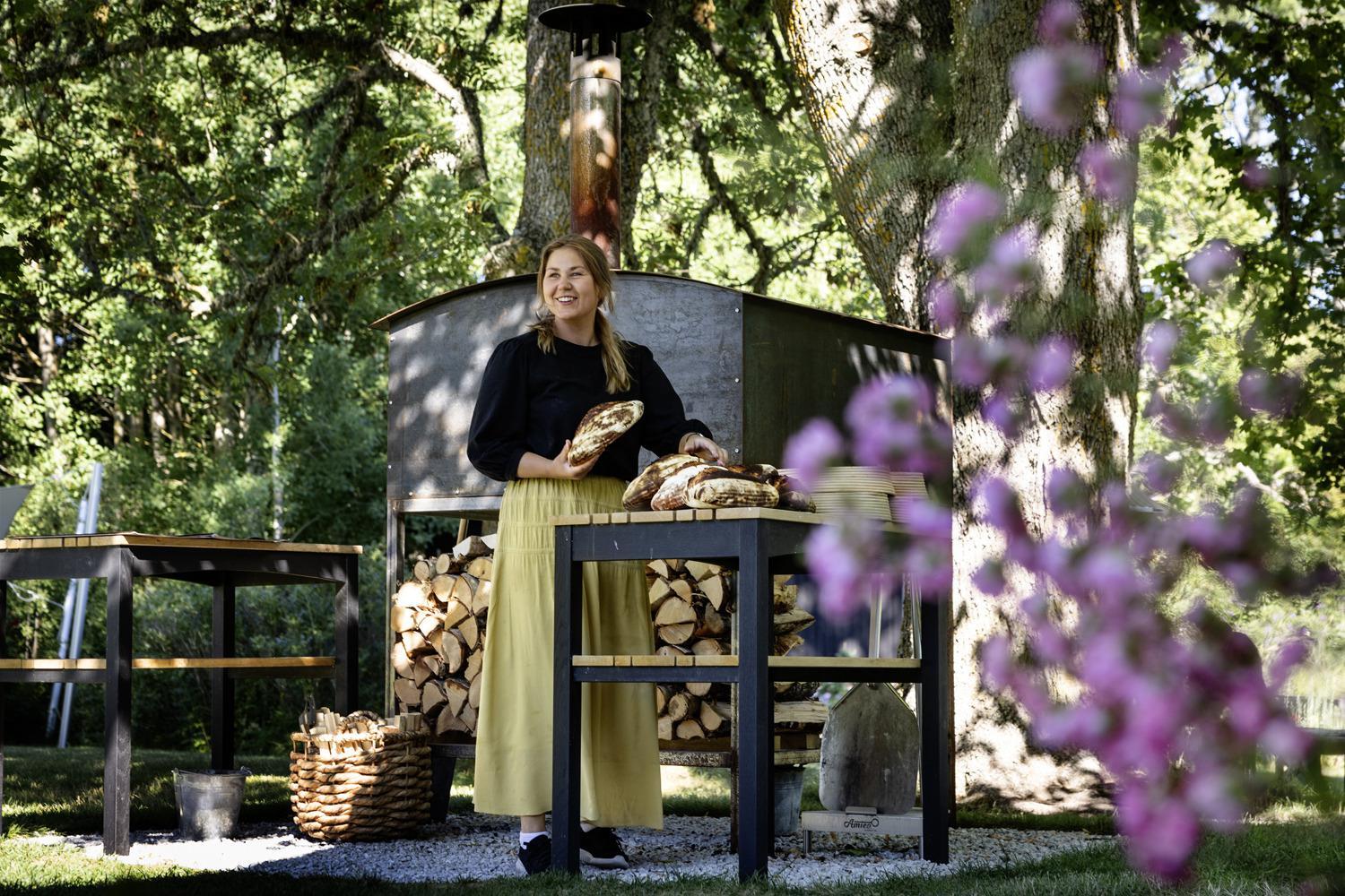 Dans un jardin pendant l'été, une femme se tient debout devant un four à bois avec du pain dans sa main.