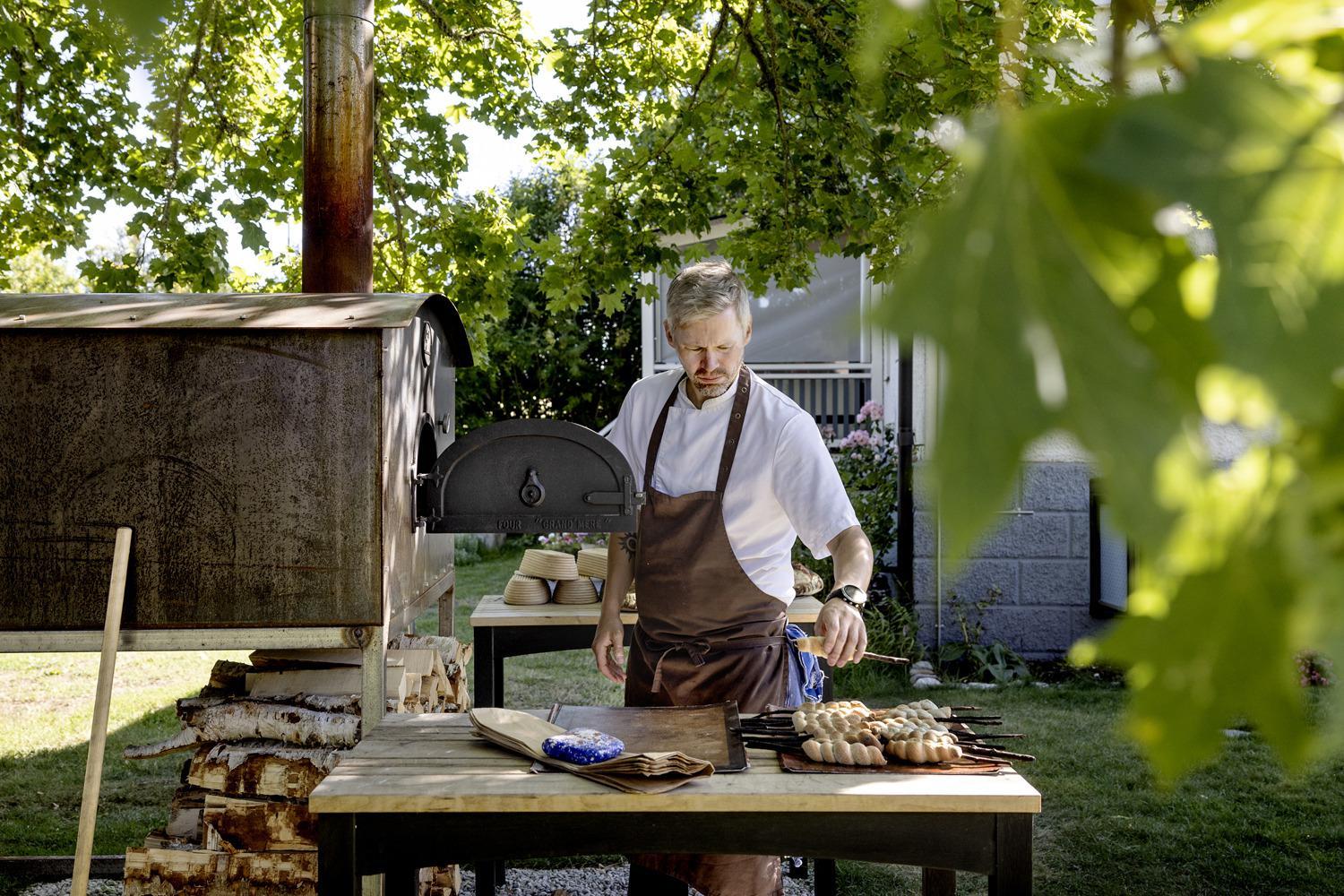 Un chef cuisinier est debout à côté d'un four à bois dans un jardin.