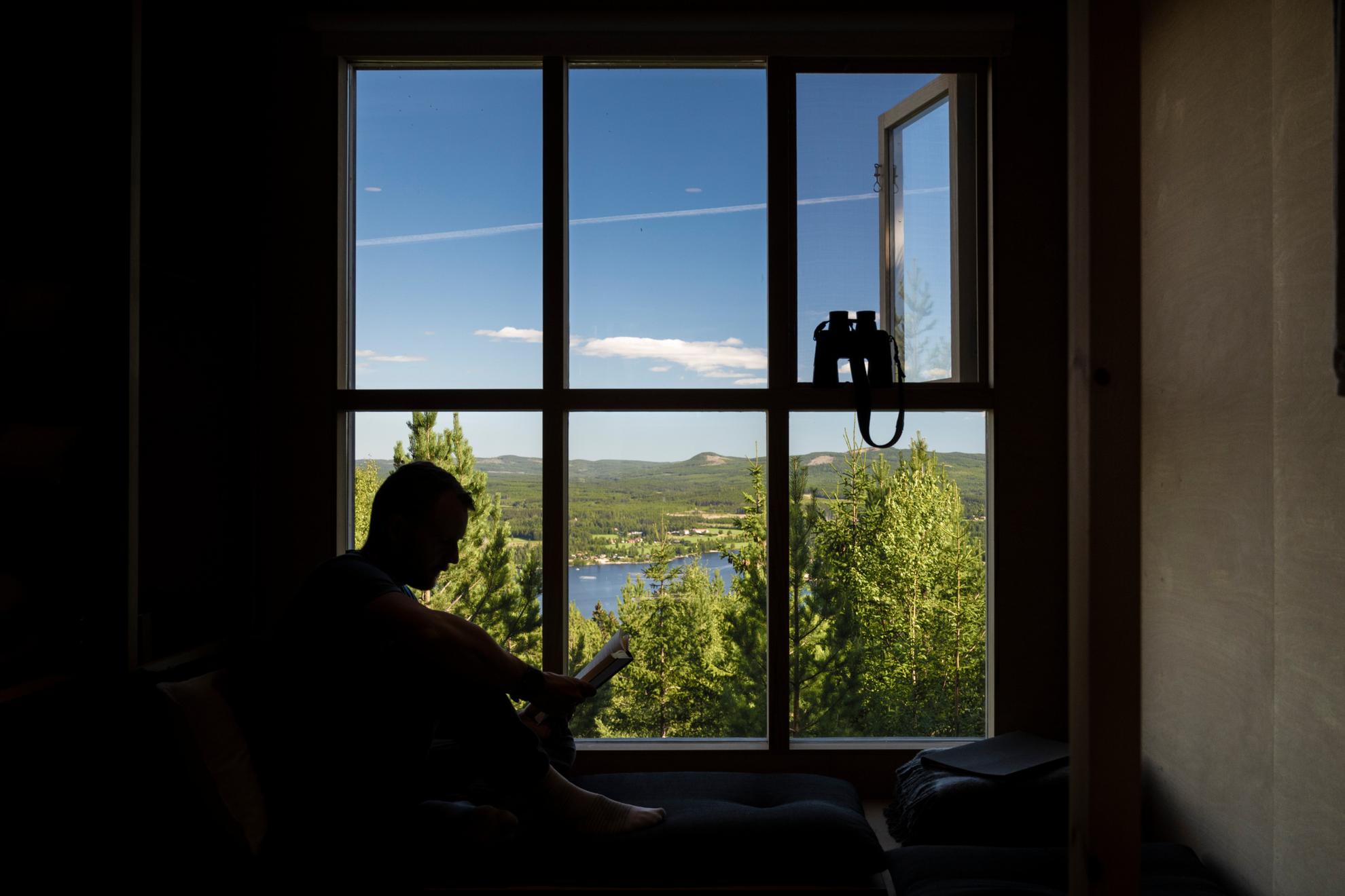 Silhouette d'un homme lisant un livre contre une grande fenêtre, regardant le ciel bleu et les montagnes du Hälsingland, avec une paire de jumelles à portée de main.
