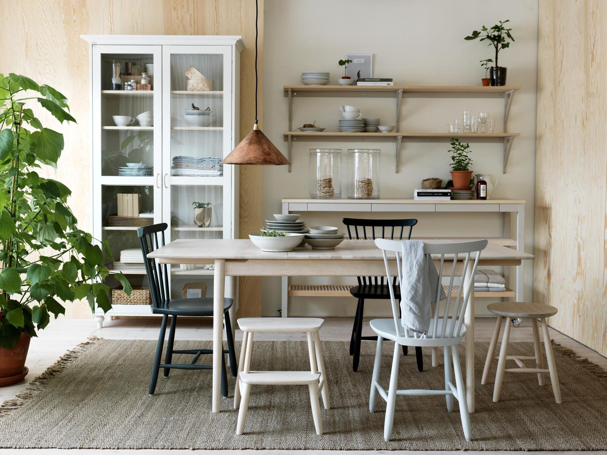 Des chaises en bois sont disposées autour d'une table en bois et des bols sont posés sur la table. Derrière, il y a une vitrine remplie et à coté des étagères de cuisine.