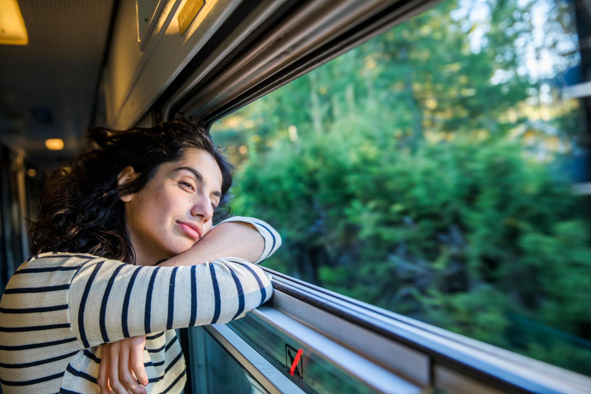 Une femme est dans un train et regarde à travers la fenêtre des arbres et de la verdure.