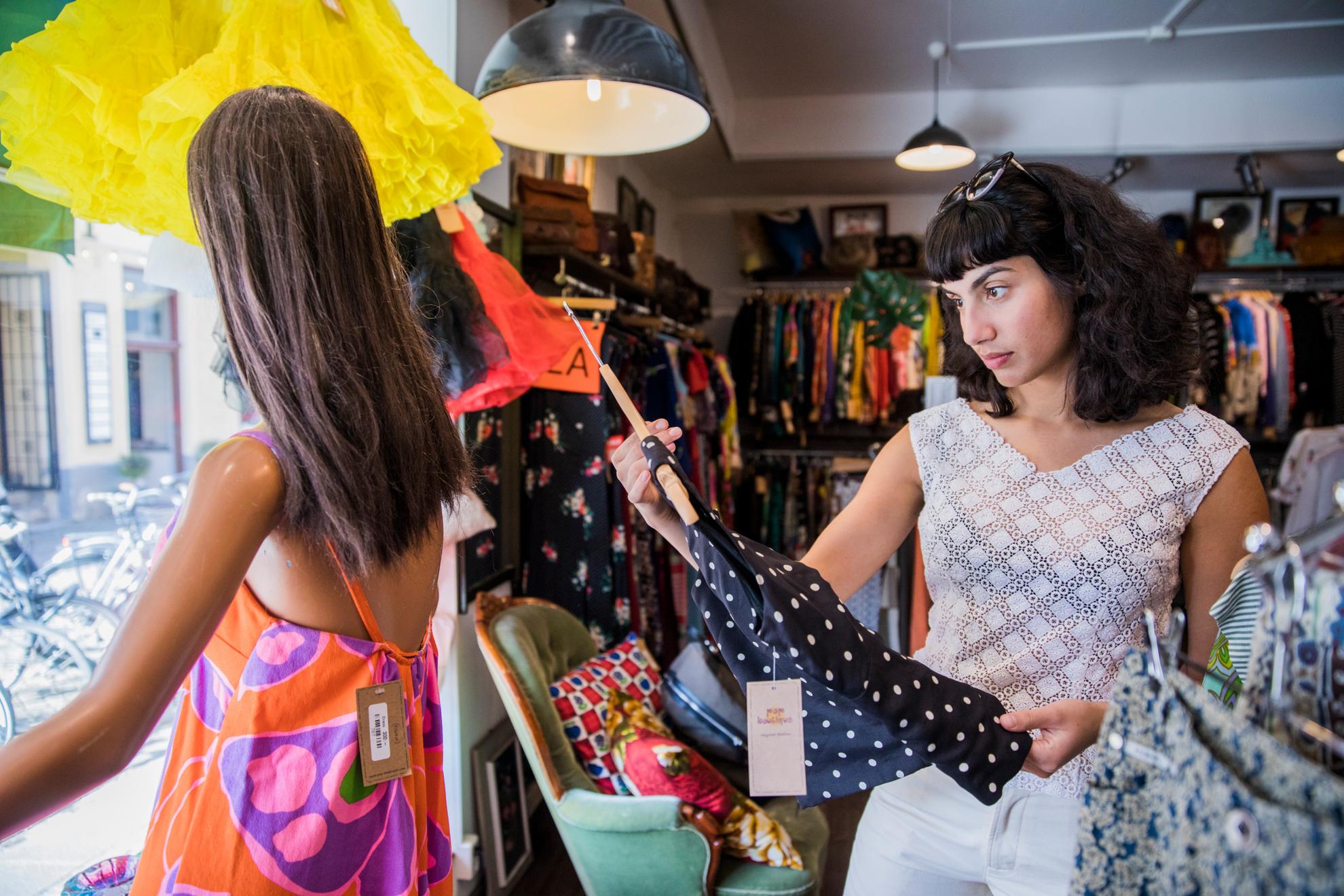 Une femme regarde un chemisier dans un magasin de vêtements vintage. À côté d'elle, se trouve un mannequin dans la vitrine, et derrière elle, on voit de nombreux vêtements.