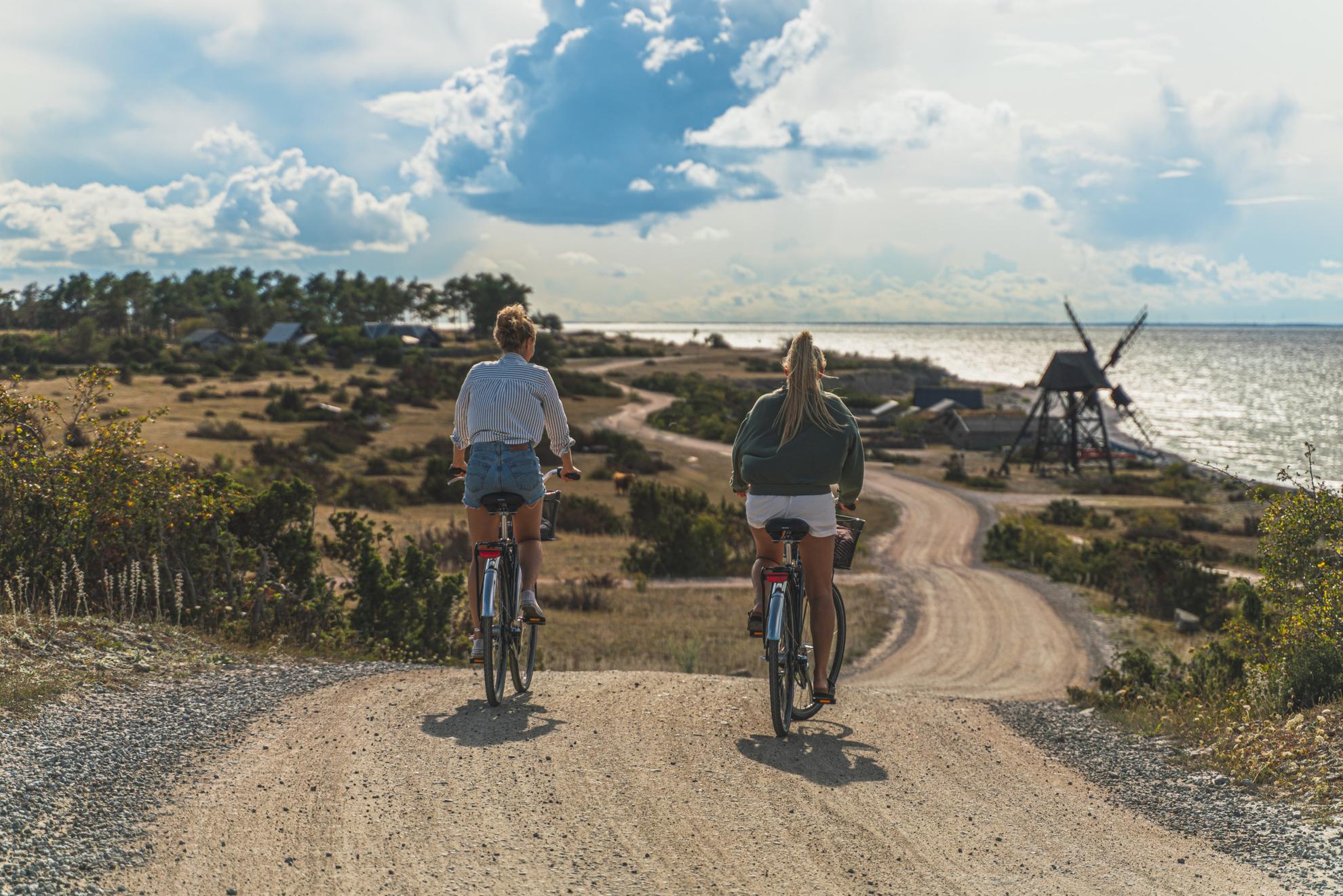 Deux femmes à vélo le long de la côte d'Öland par une belle journée ensoleillée, avec un moulin à vent en arrière-plan.