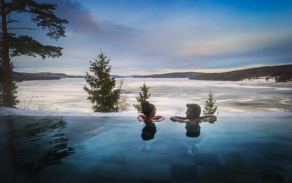 Deux personnes se détendent dans la piscine extérieure de l'Orbaden Spa & Resort à Hälsingland, en regardant la vue sur l'eau recouverte de neige.