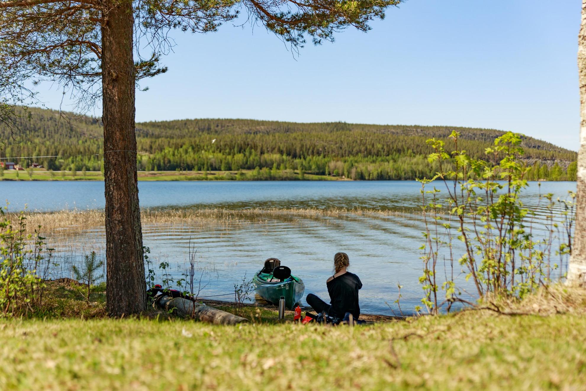 Une femme est assise sur la rive d'un lac avec son canoë au bord de l'eau. On aperçoit derrière le lac, une vaste forêt.