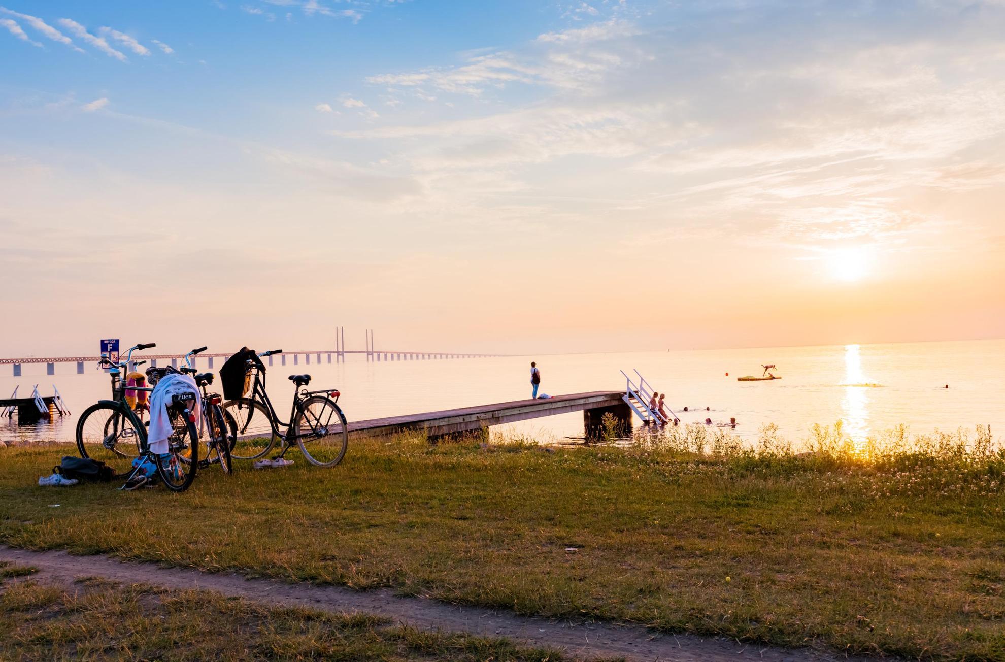 Coucher de soleil sur une plage avec une jetée. Deux vélos sont garés sur l'herbe au-dessus de la plage et plusieurs personnes se baignent dans la mer. Le pont de l'Öresund en arrière-plan.