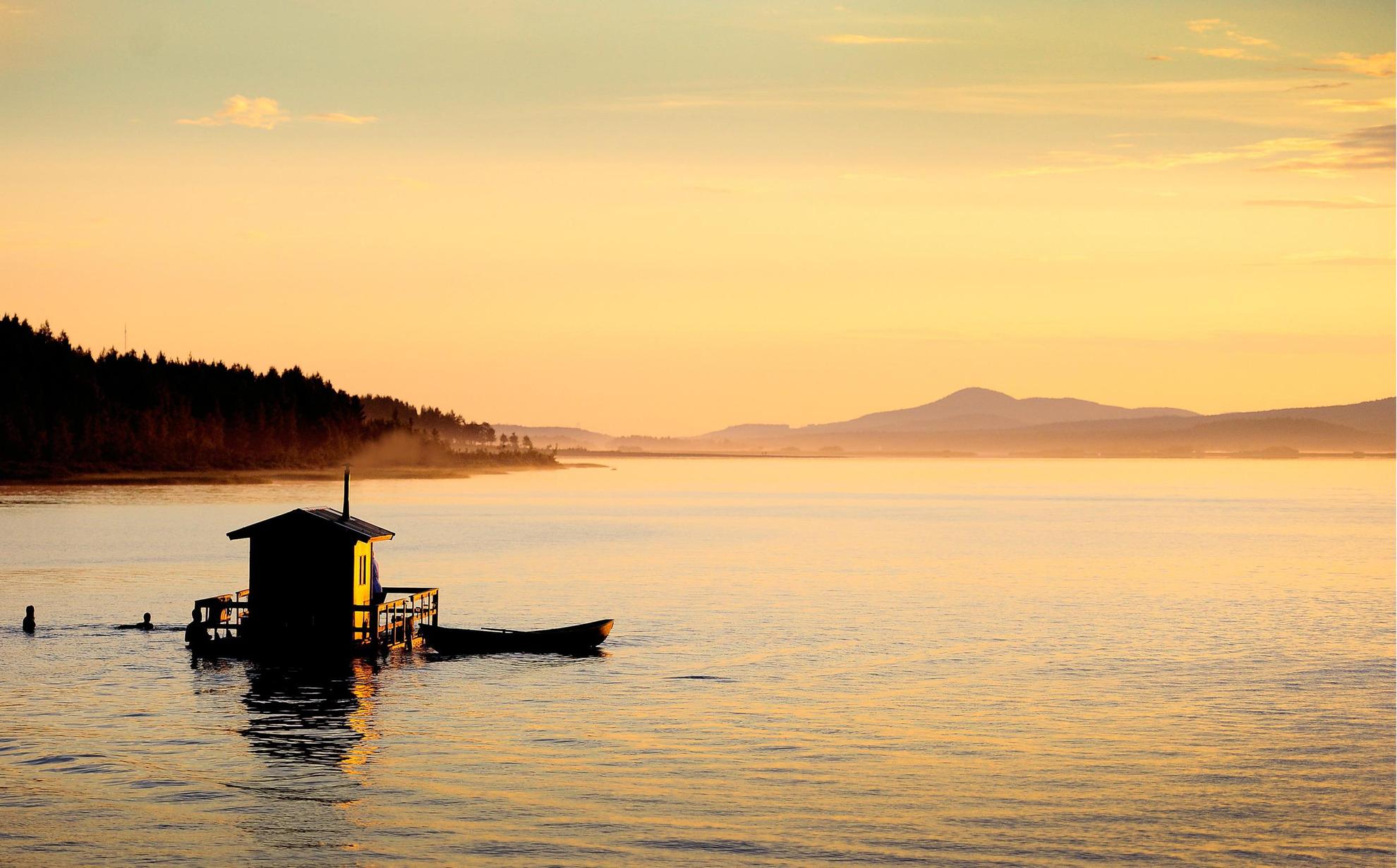 Un bateau-sauna sur un lac au coucher du soleil. Les gens se baignent près du sauna. Une barque est amarrée au bateau-sauna.