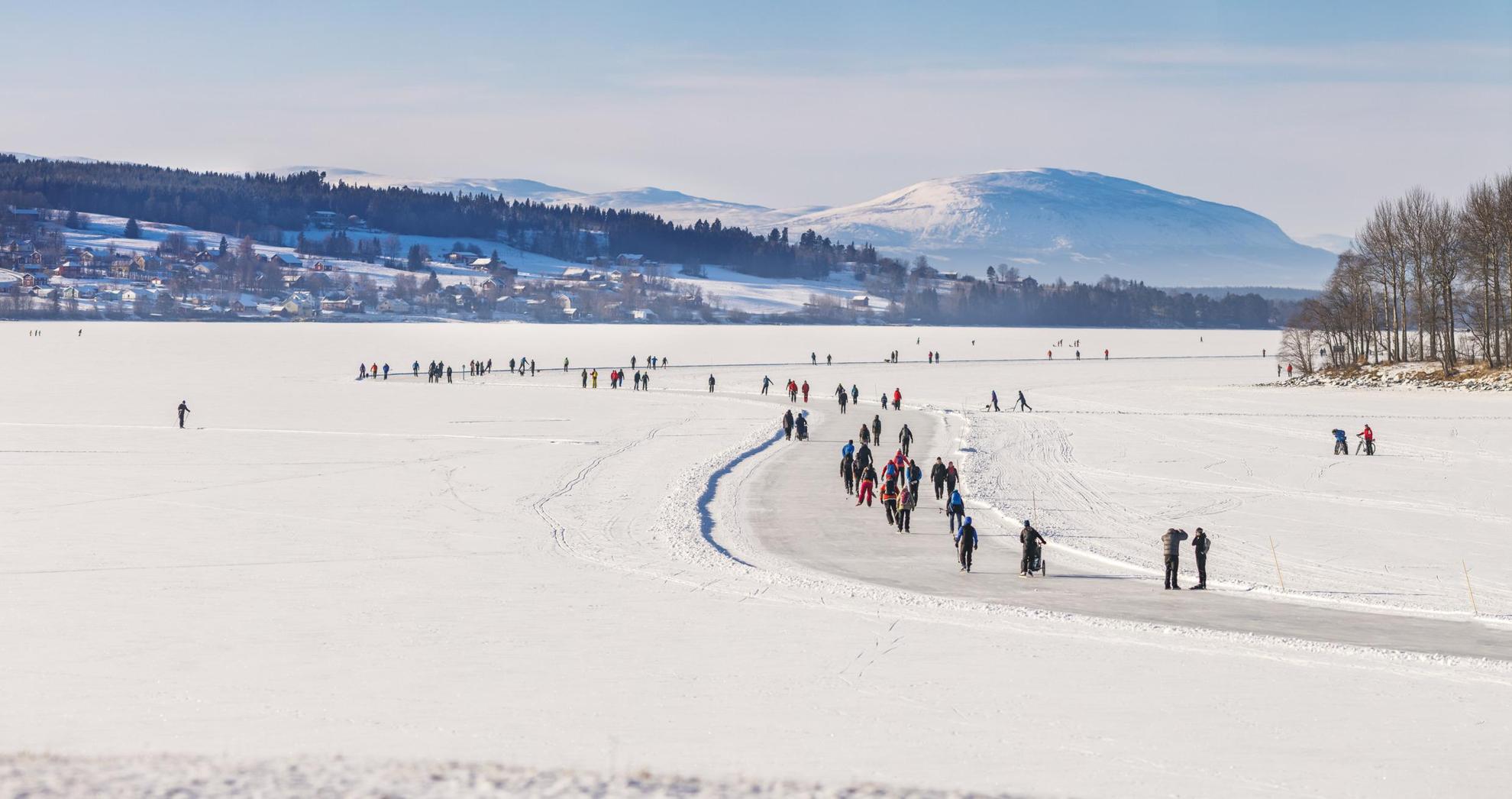 Plusieurs personnes font du patin à glace sur des pistes préparées sur un lac gelé recouvert de neige. On voit en arrière plan, des maisons, des arbres et des montagnes.