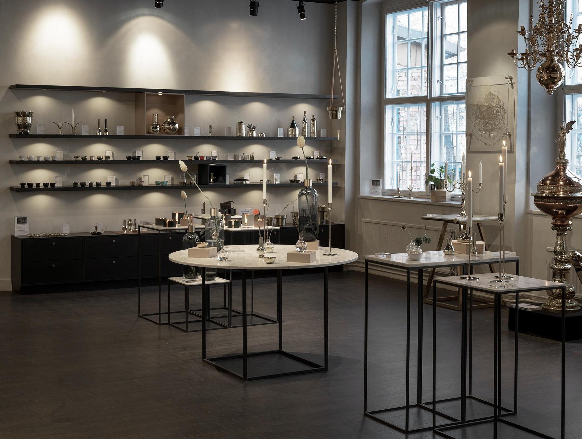 Photo de l'intérieur de la boutique de Skultuna Messingsbruk avec des étagères noires et des tables blanches où sont exposés divers produits tels que des vases et des bougeoirs.