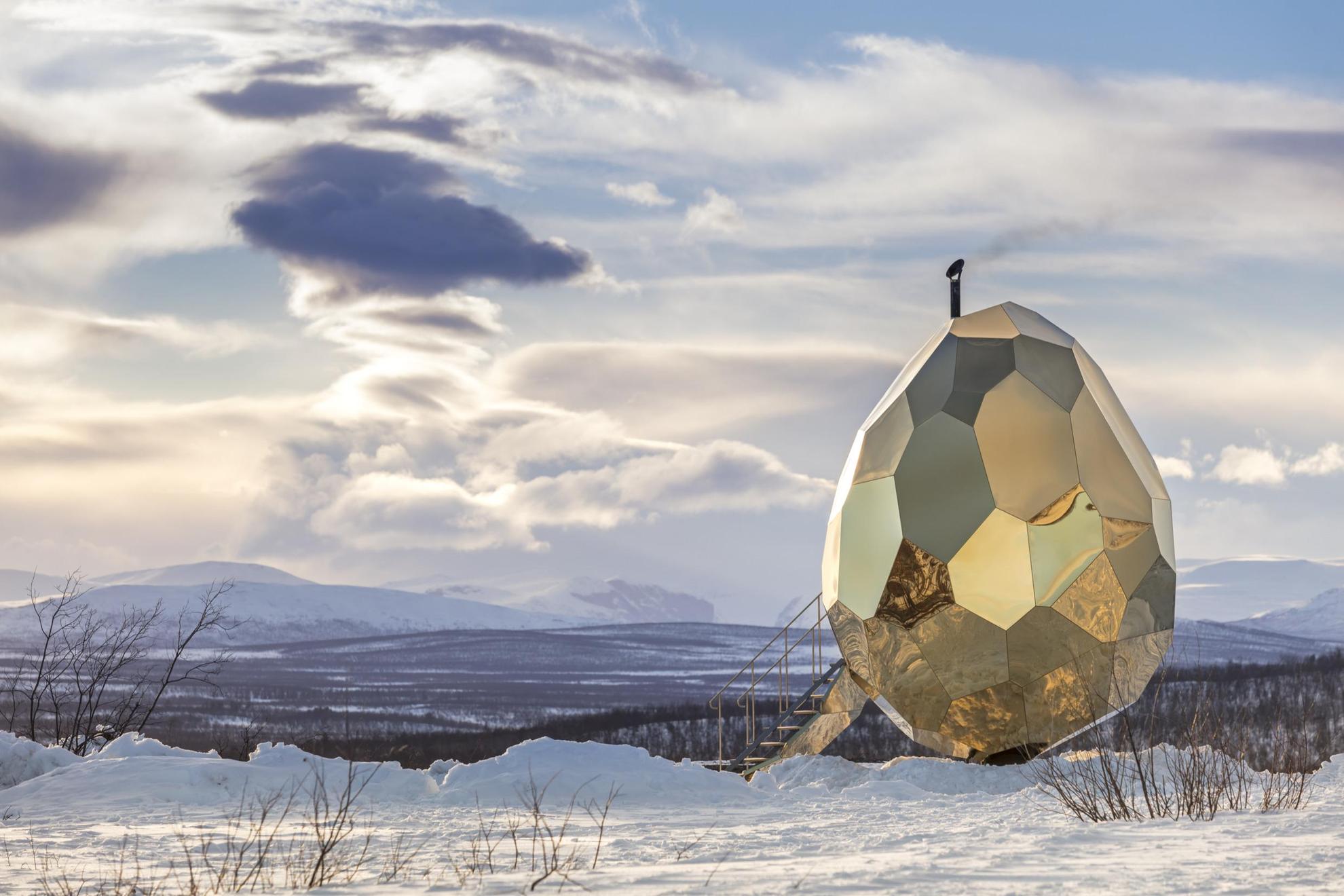 Un sauna au feu de bois en forme d'œuf avec des morceaux de coquille réfléchissants est installé dans un paysage hivernal arctique de Kiruna avec, en arrière-plan, une vue panoramique sur la vallée et les montagnes enneigées au loin.