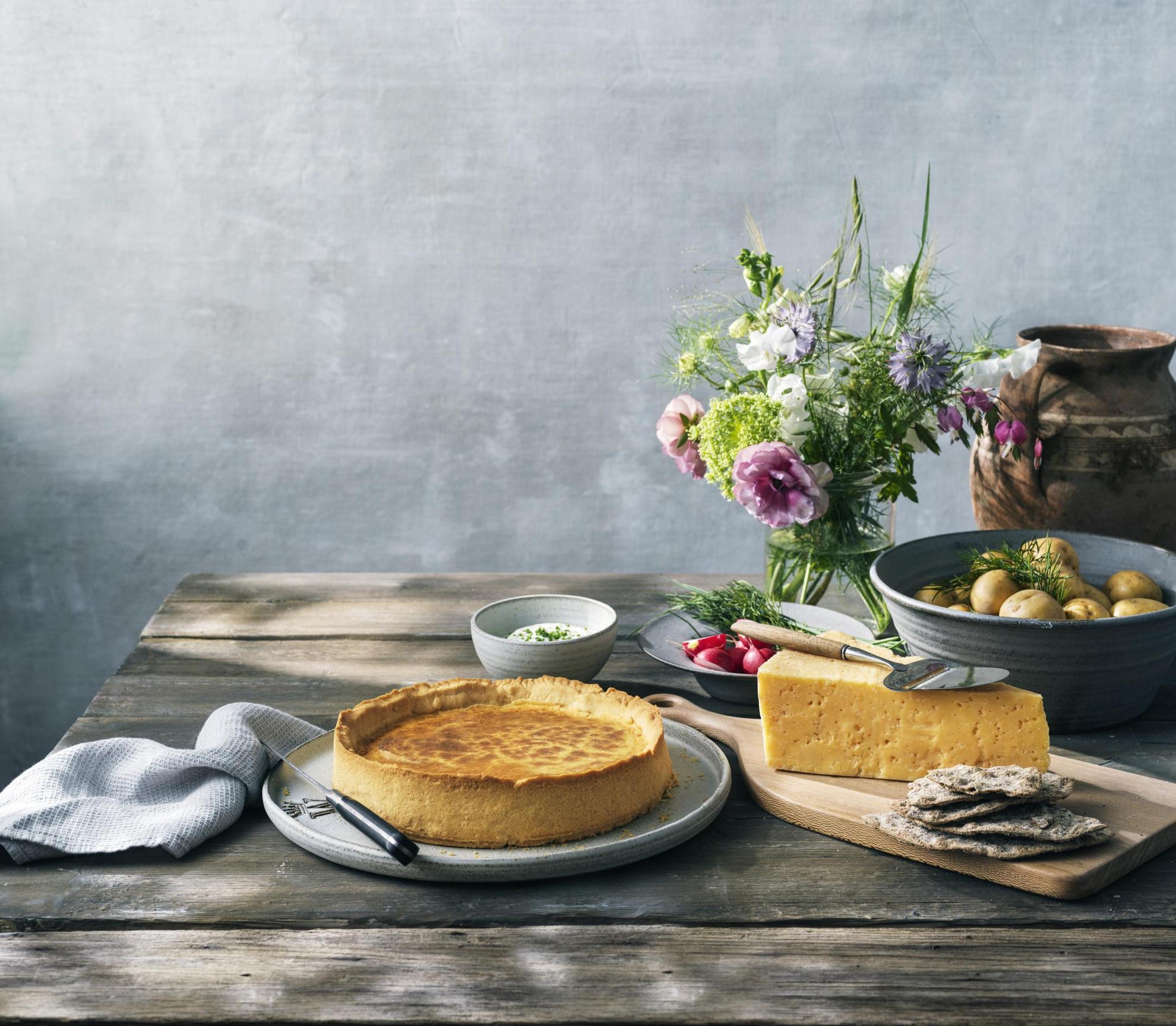 Une table en bois avec une tarte Västerbottenpaj, du fromage Västerbotten, un bol de pommes de terre et un vase de fleurs.