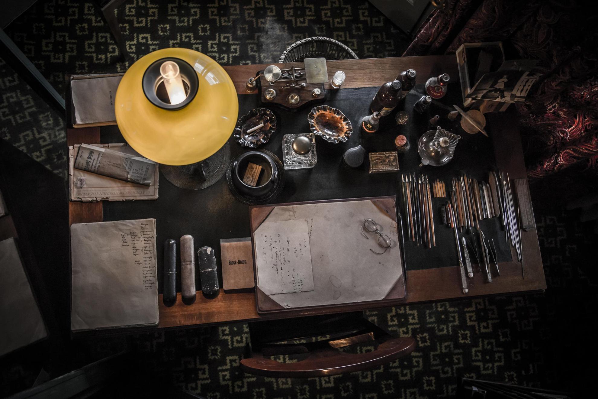 Un vieux bureau en bois, avec des crayons alignés les uns à côte des autres, du papier, des notes, des verres et une vieille lampe.