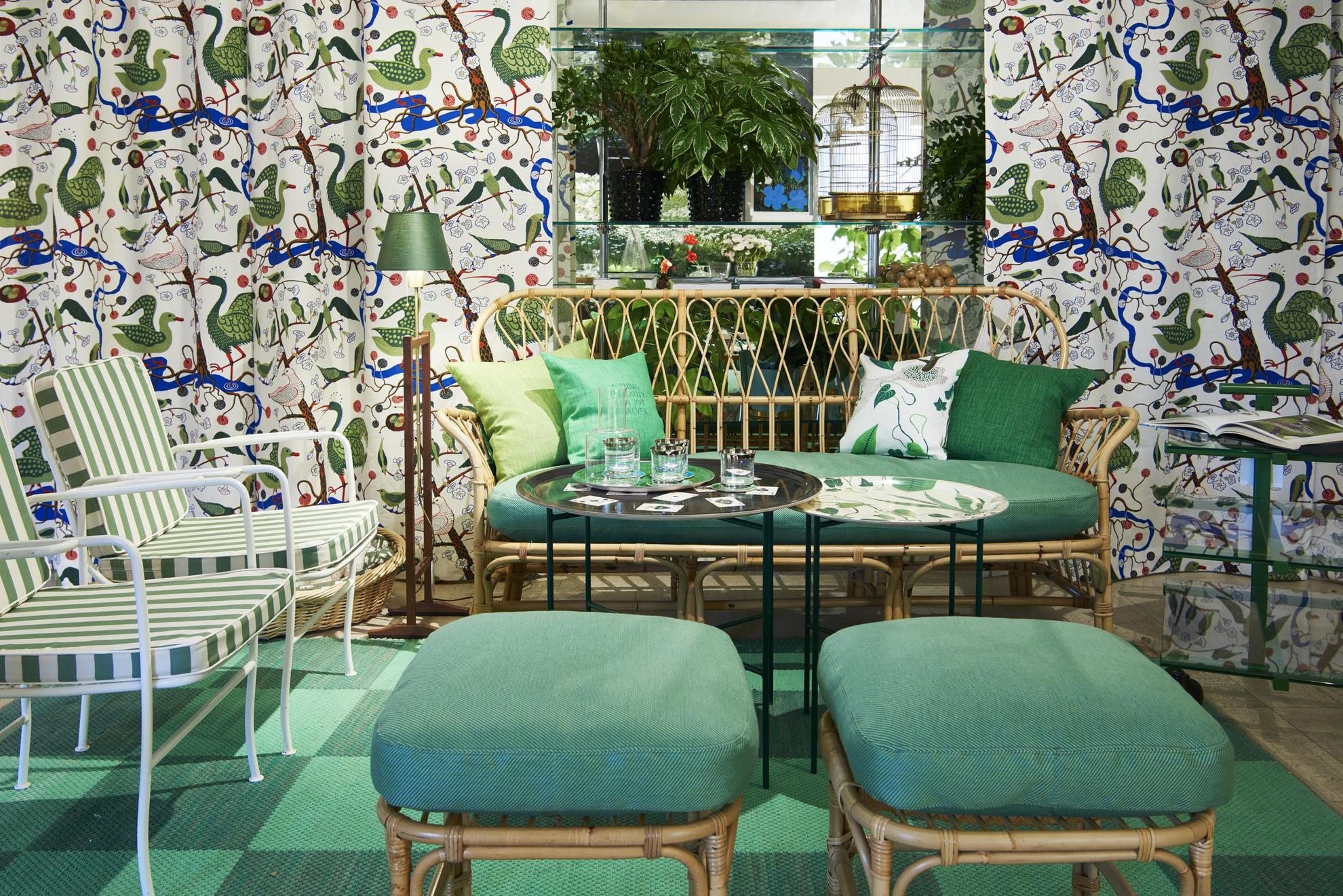 Un environnement vert dans un magasin de design. Un canapé, deux fauteuils et deux poufs sont disposés autour d'une table. Derrière les meubles sont installés des rideaux colorés avec des oiseaux en vert.