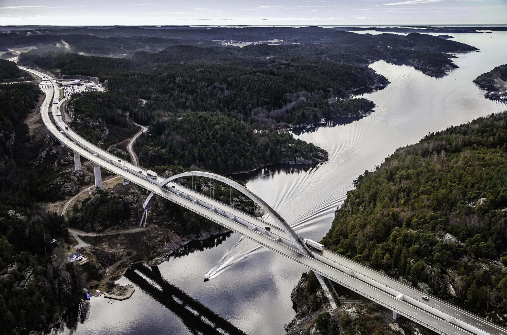 The Svinesund Bridge has traffic on it, and underneath a motorboat is racing. The landscape on both sides of the bridge is hilly.