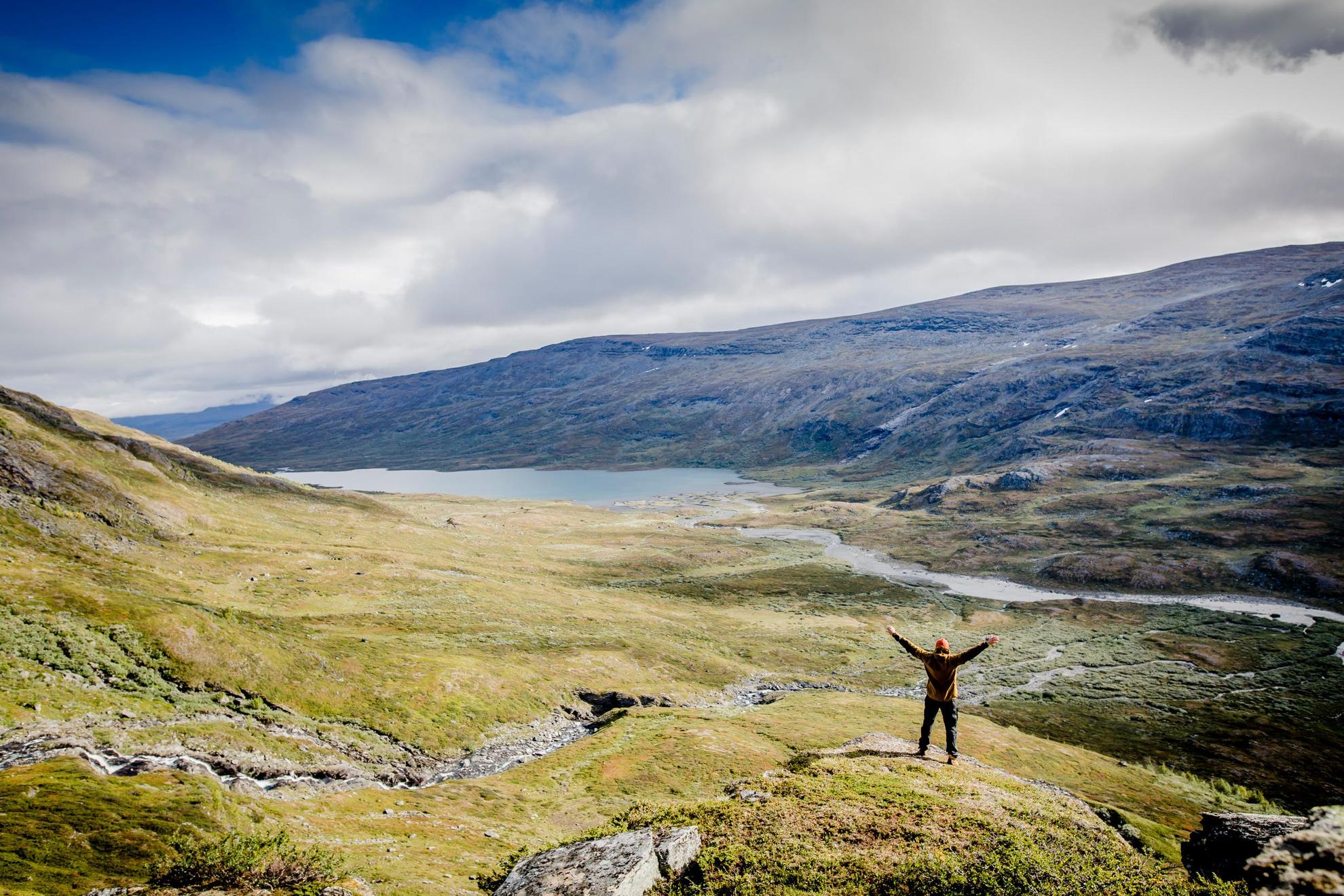 Une personne lève les bras vers le ciel. Elle se tient devant un paysage scénique composé d'une vallée traversée par un lac et une rivière et bordée de montagnes.