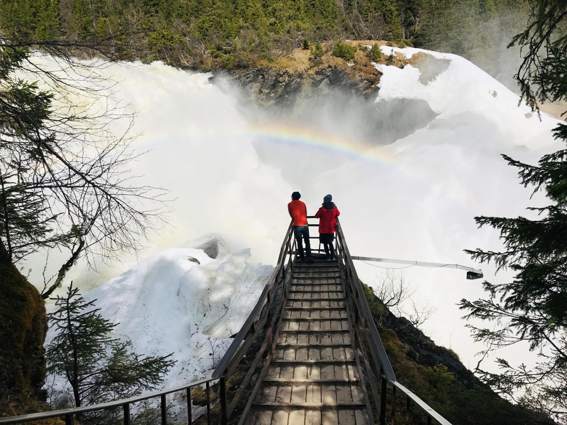 Deux personnes sont en train d'admirer une grande chute d'eau depuis un ponton surélevé. L'eau de la cascade crée un arc-en-ciel.