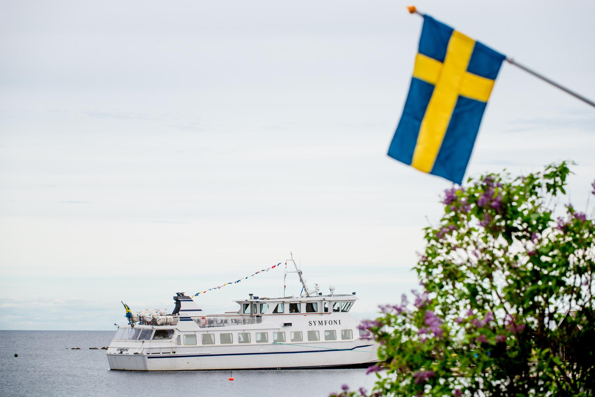 Un bateau nommé Symfoni flotte sur l'eau, avec un drapeau suédois et des fleurs violettes au premier plan.