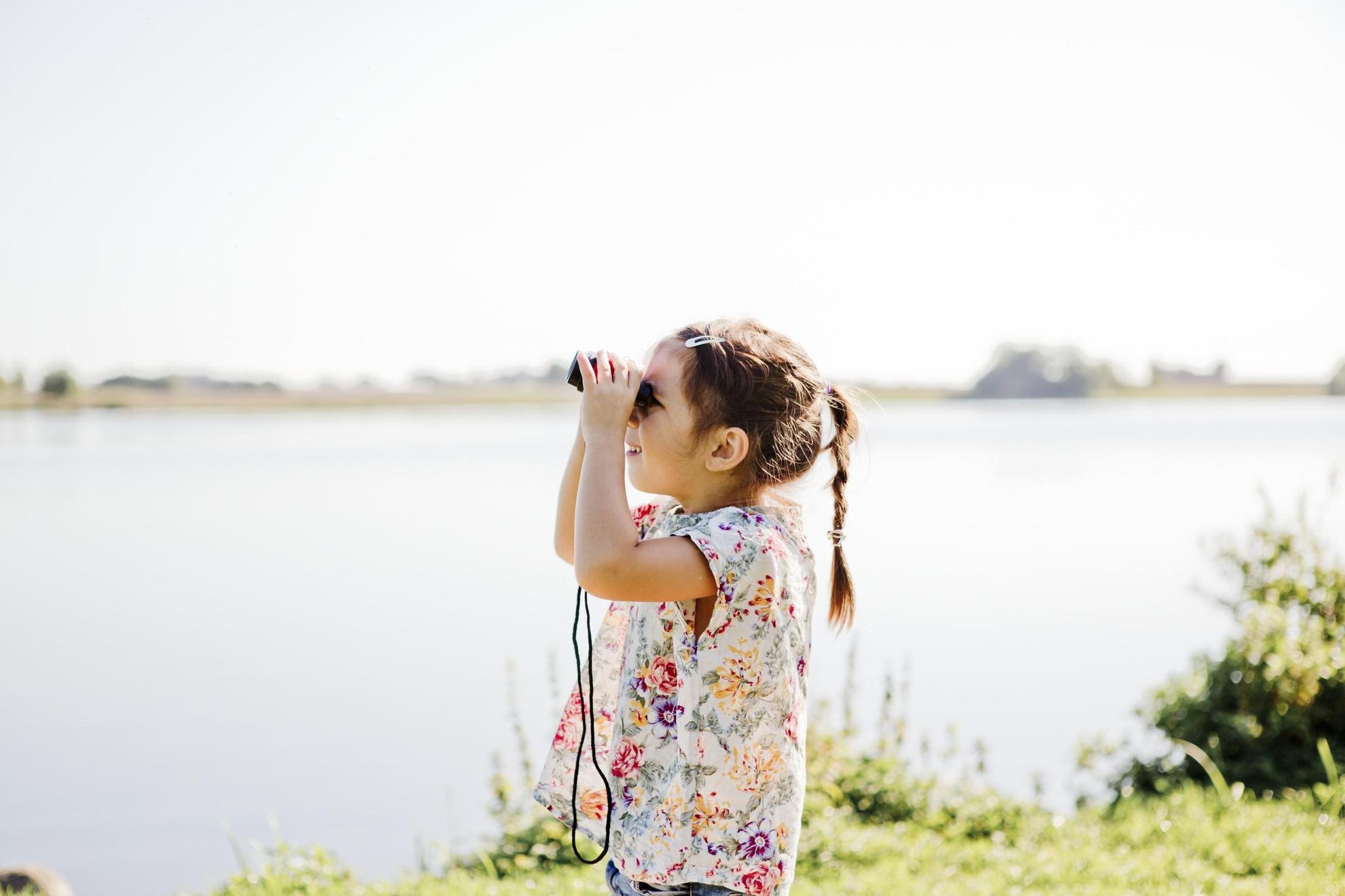 A girl by a lake is looking through binoculars.