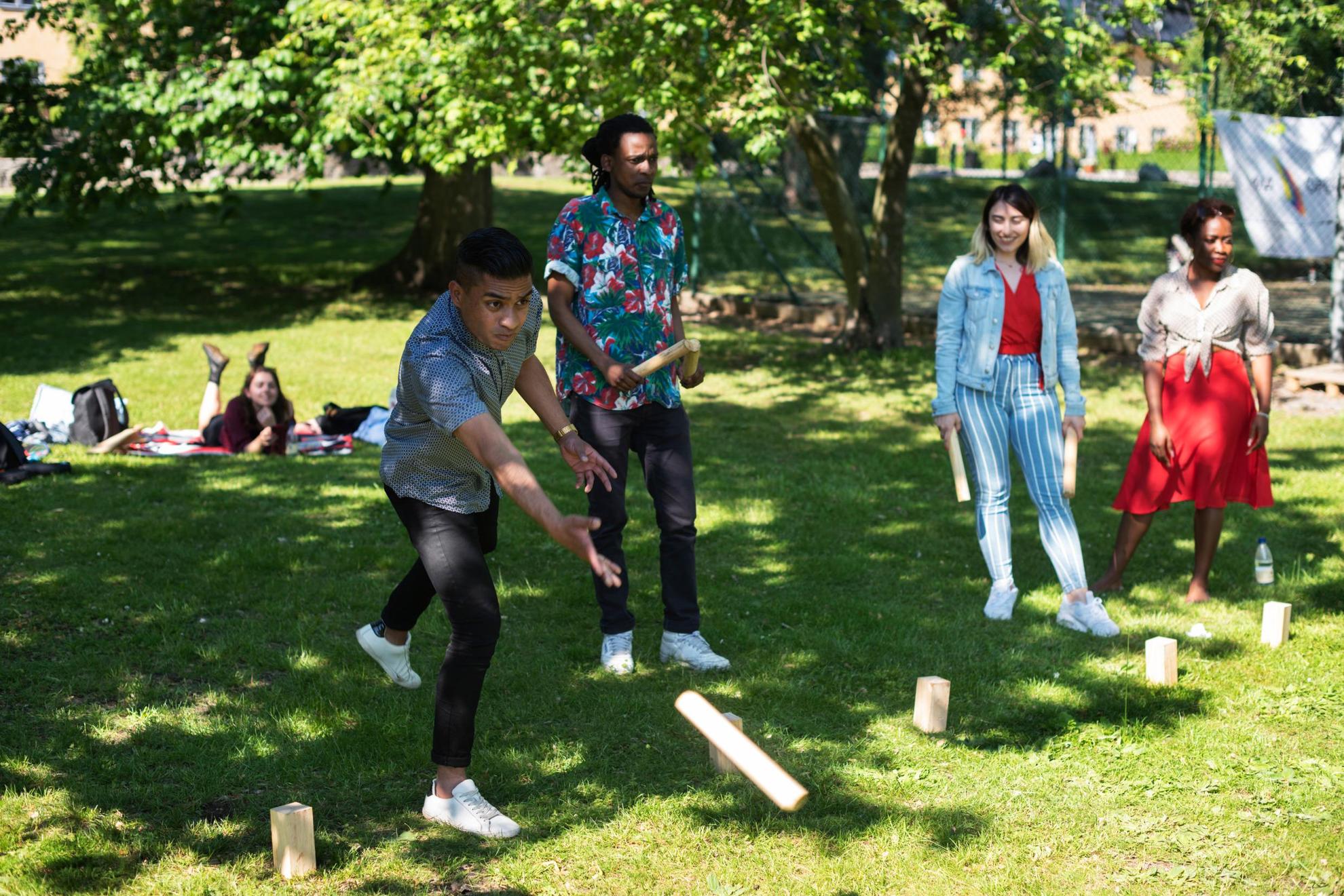 Des personnes sont dans un parc et lancent des bâtons en bois sur des blocs de bois posés dans l'herbe.
