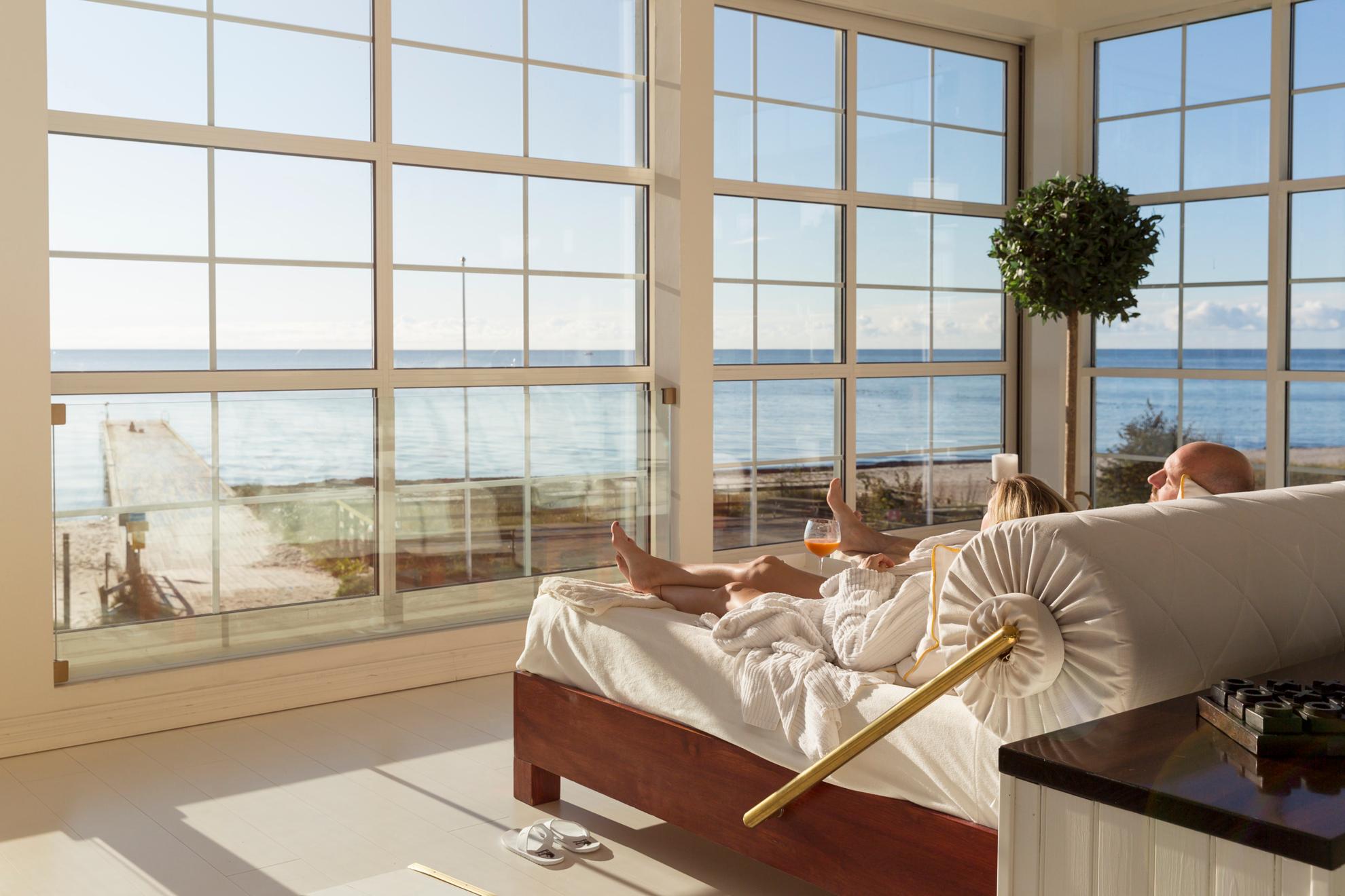 Un couple est allongé dans un grand lit à côté de grandes fenêtres donnant sur la plage et la mer.