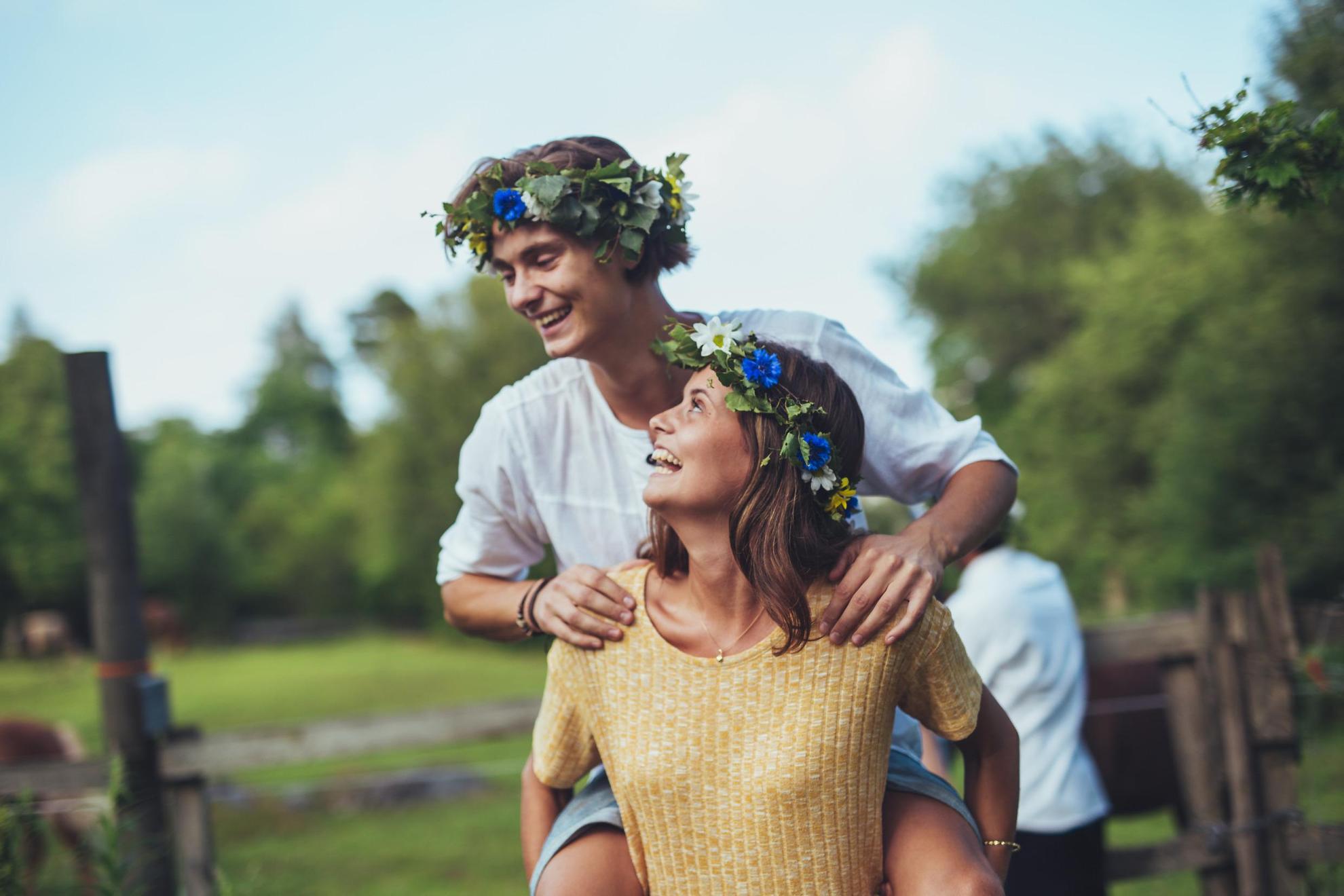 Une femme porte un homme sur son dos dans la nature. Les deux portent des couronnes de fleurs de midsommar dans leurs cheveux.
