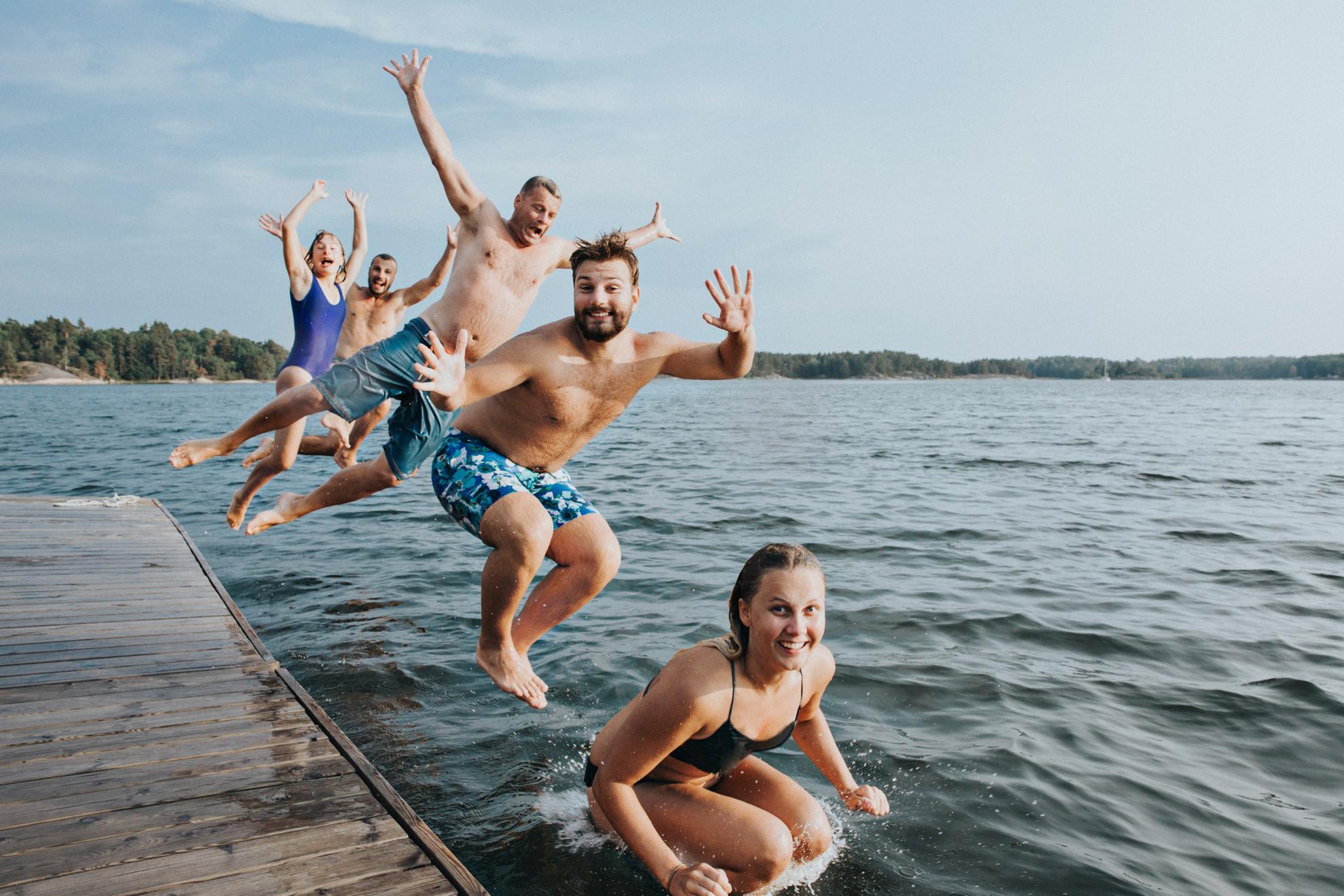 Cinq jeunes sont en maillot de bain et sautent dans un lac depuis un ponton en bois.